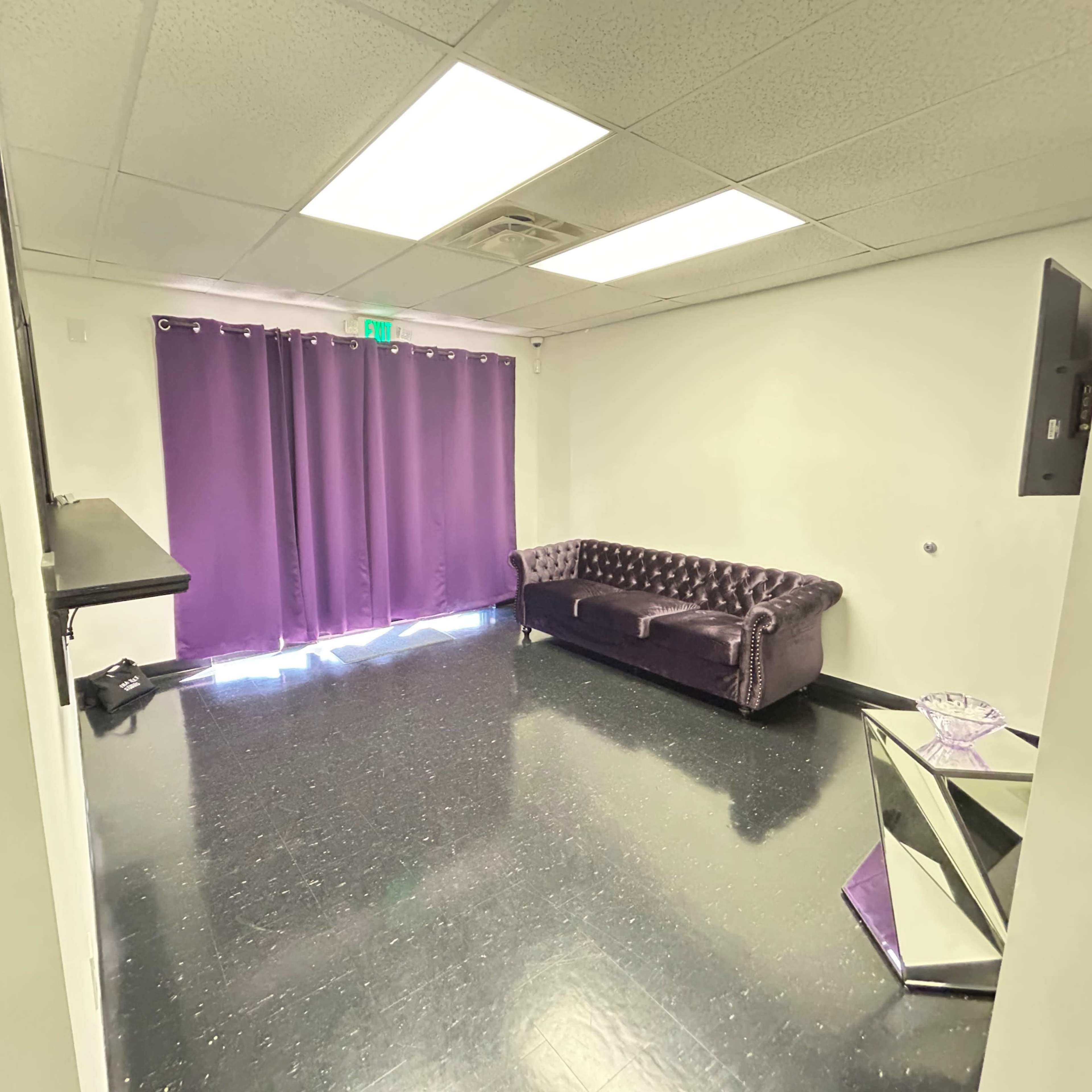 A sparsely furnished room with a purple curtain, a dark sofa, and a reflective coffee table.