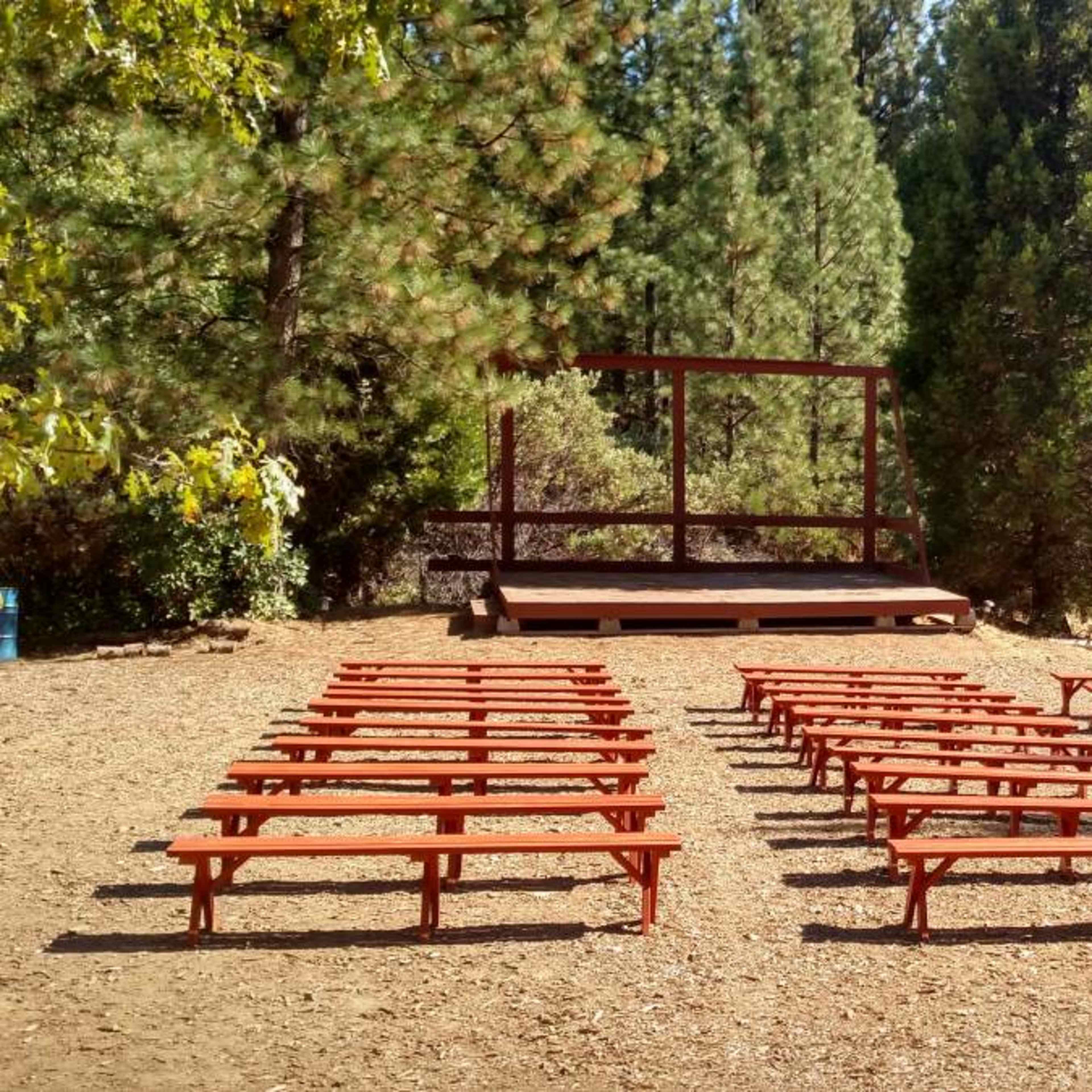 A clearing with rows of wooden benches facing a small stage surrounded by trees.