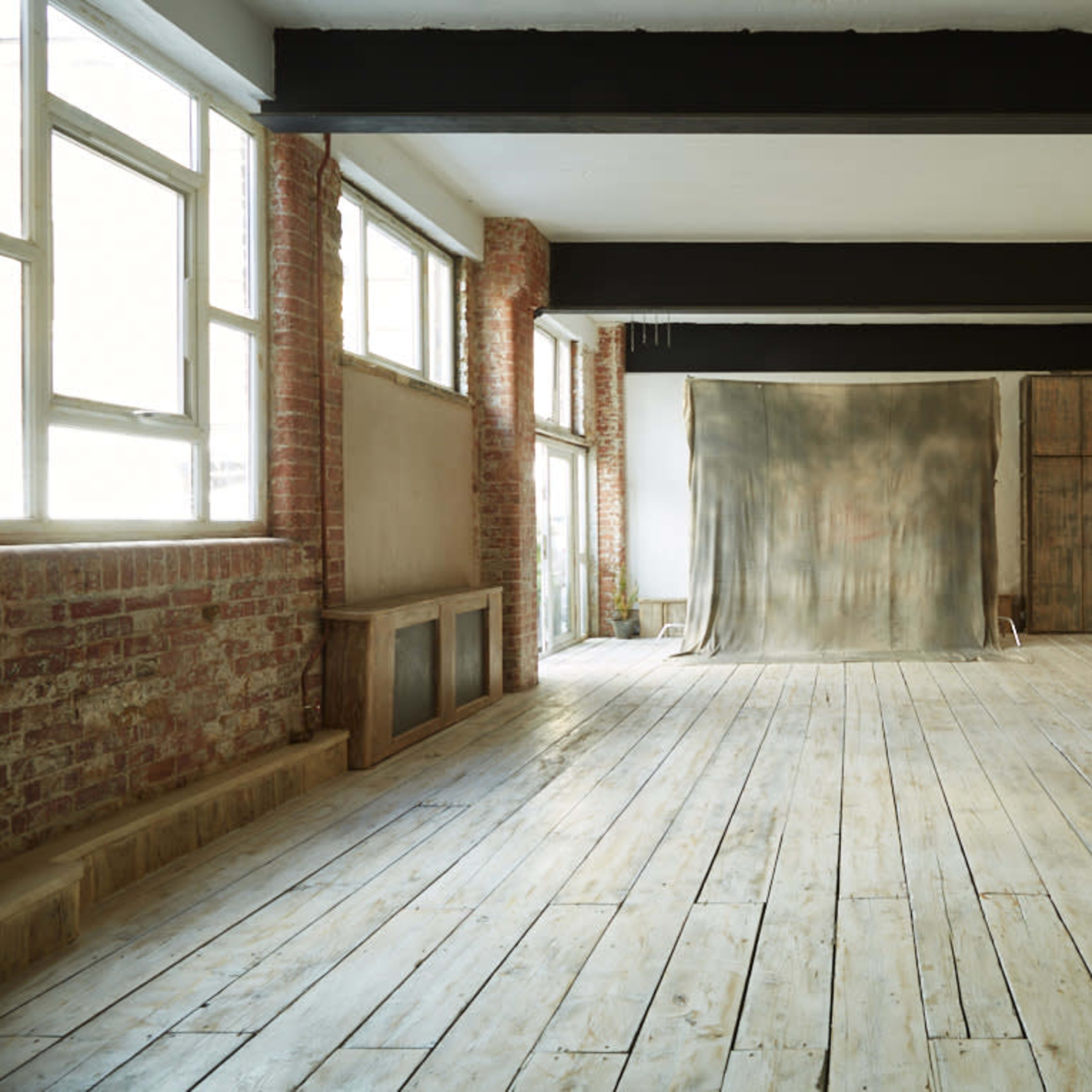 An empty studio space with large windows, exposed brick walls, and a wooden floor, featuring a plain backdrop in the center.