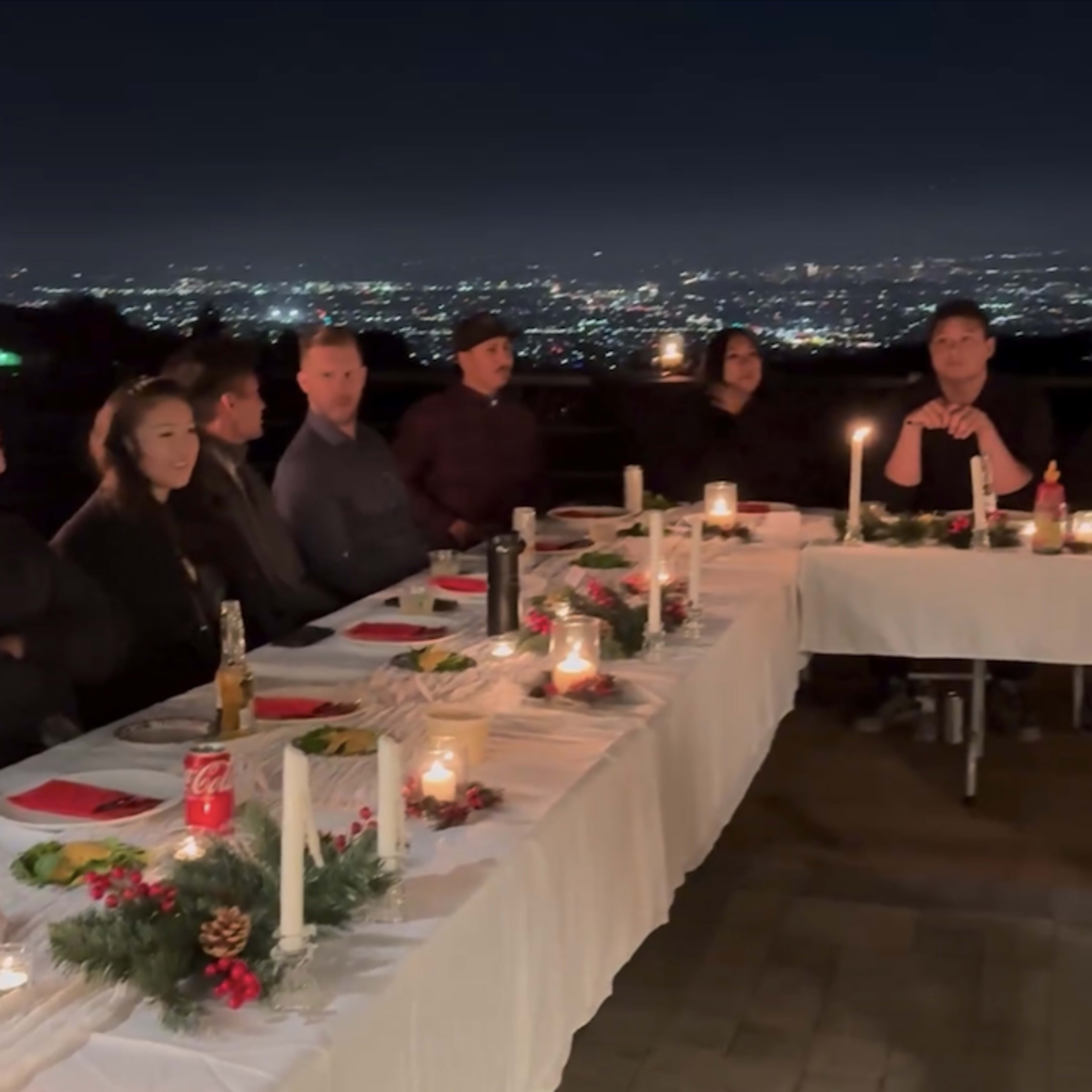 A group of people is seated at a long table decorated with candles and festive items, overlooking a city skyline at night.