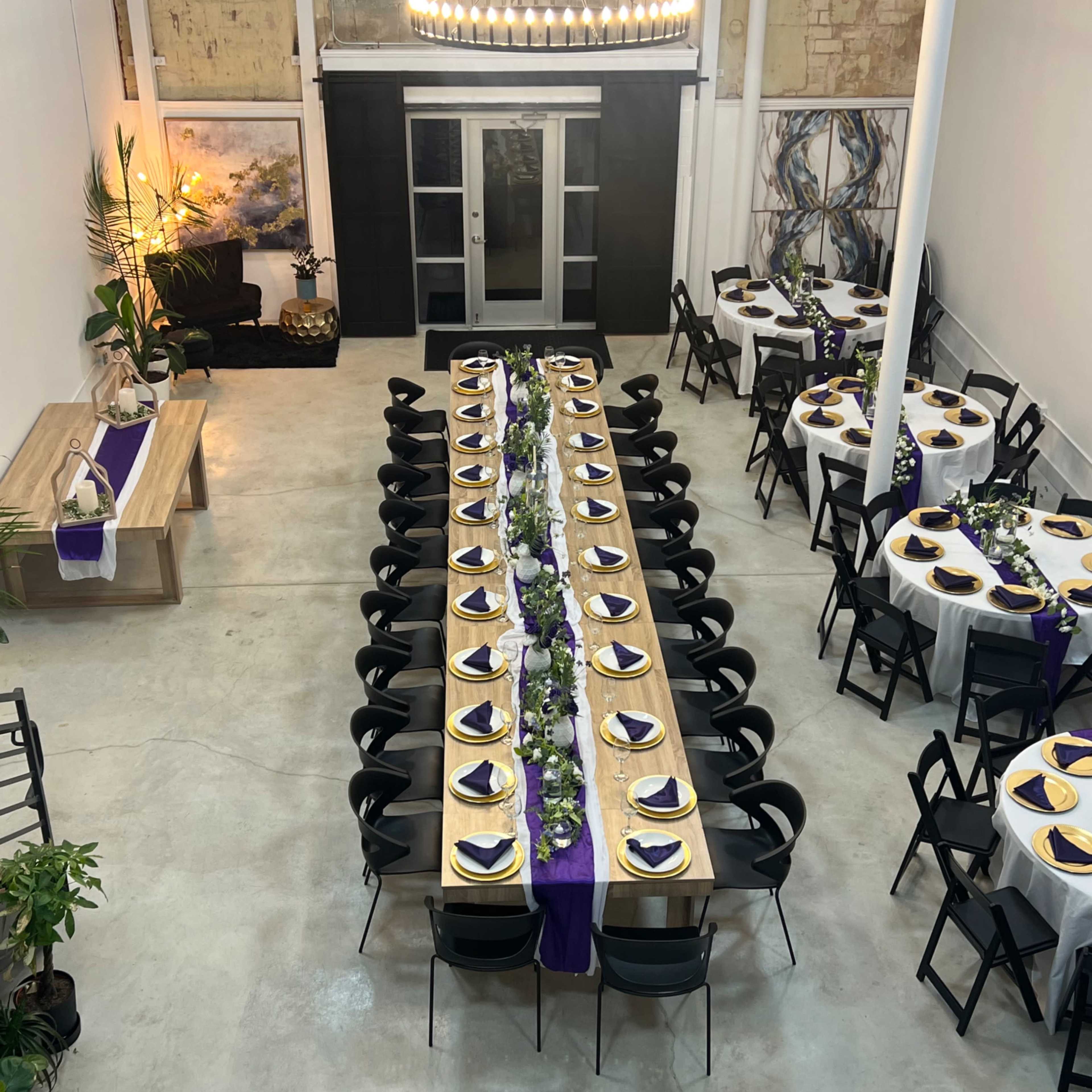 A large dining area features two long tables set with white tablecloths and purple accents, arranged for a formal event in a spacious room with exposed brick walls.