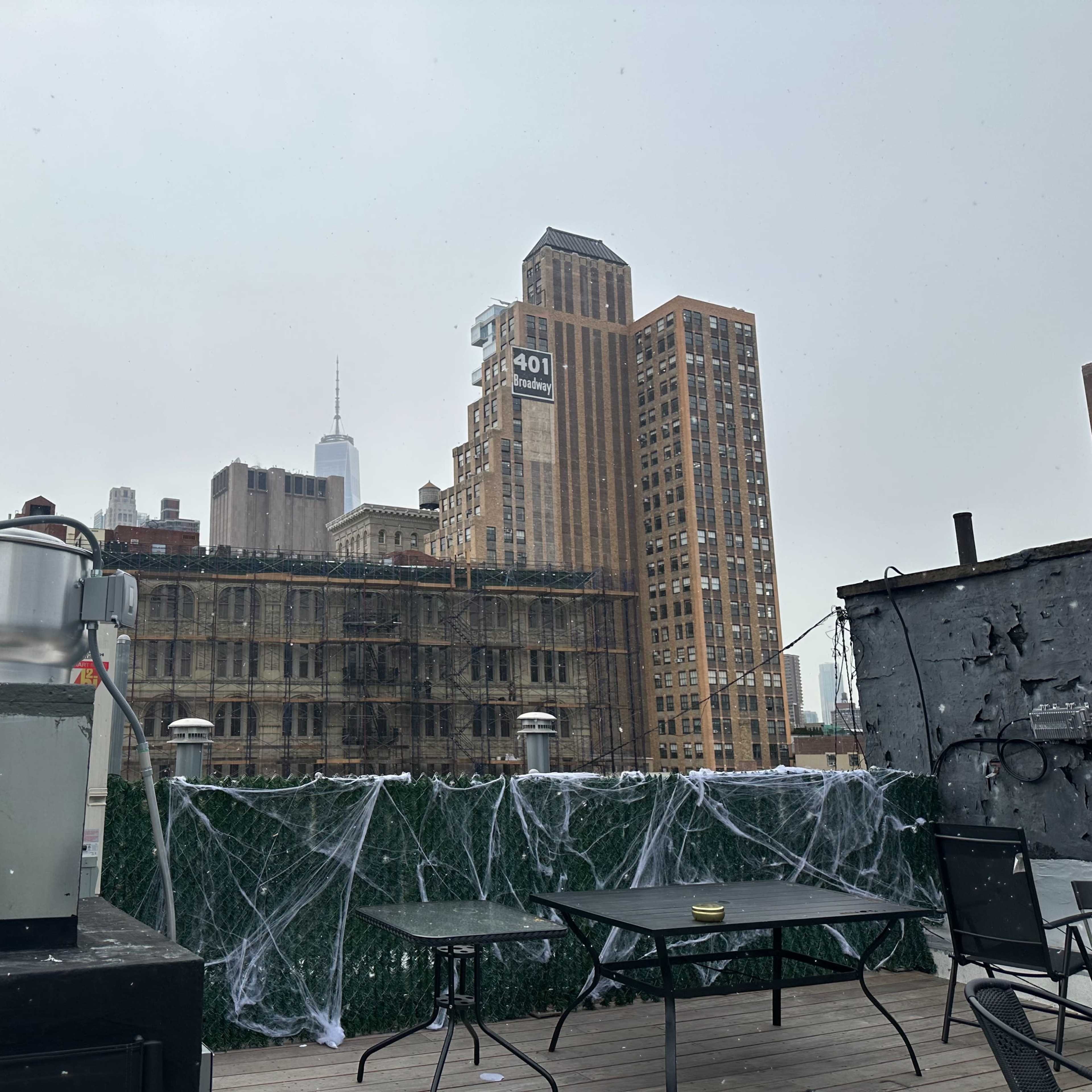 A rooftop scene features a stainless steel grill, tables, and buildings, including a tall structure with a spire in the background, all partially obscured by a foggy sky.