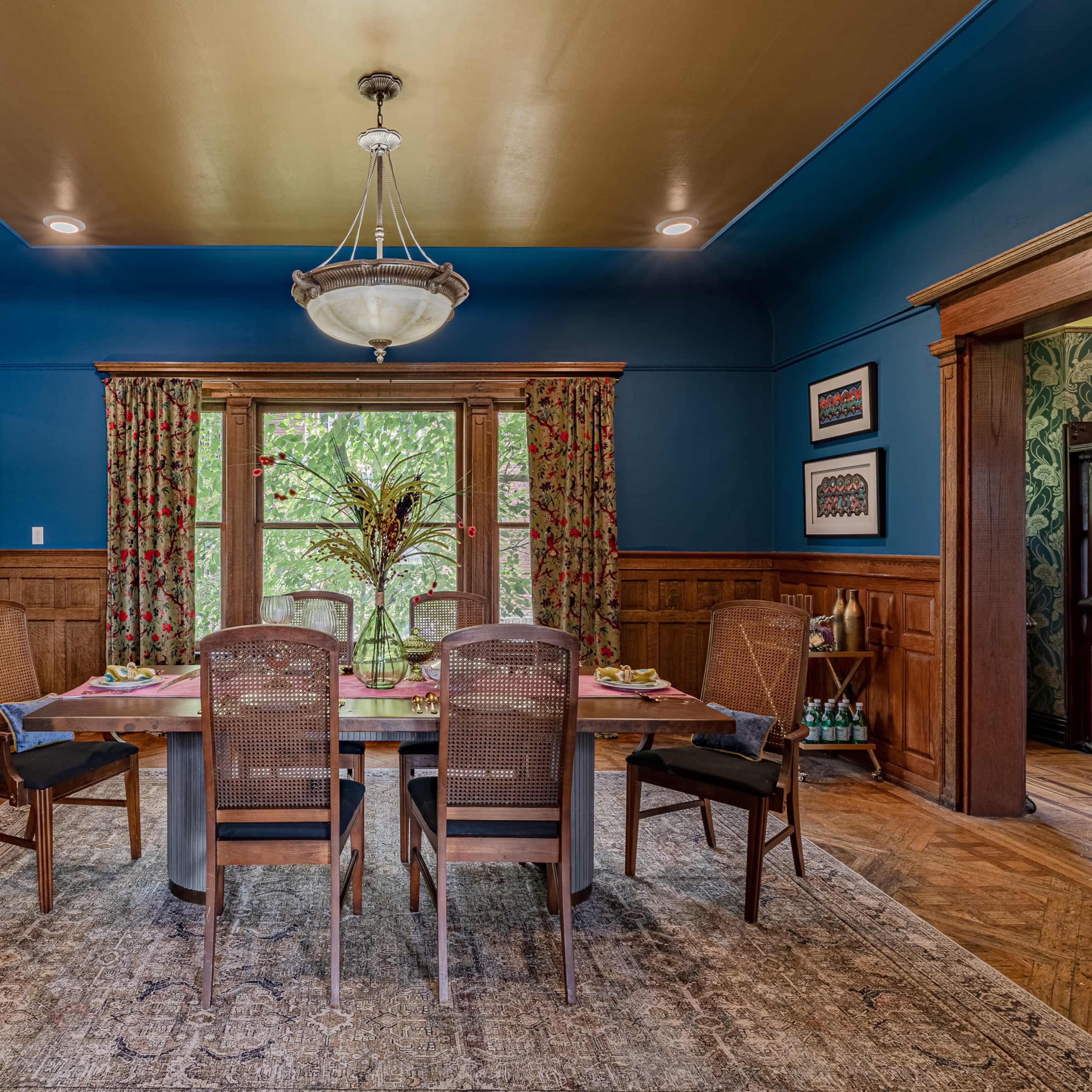 A dining room features a wooden table set for a meal, surrounded by four chairs, with decorative curtains, a chandelier, and richly stained wood paneling.