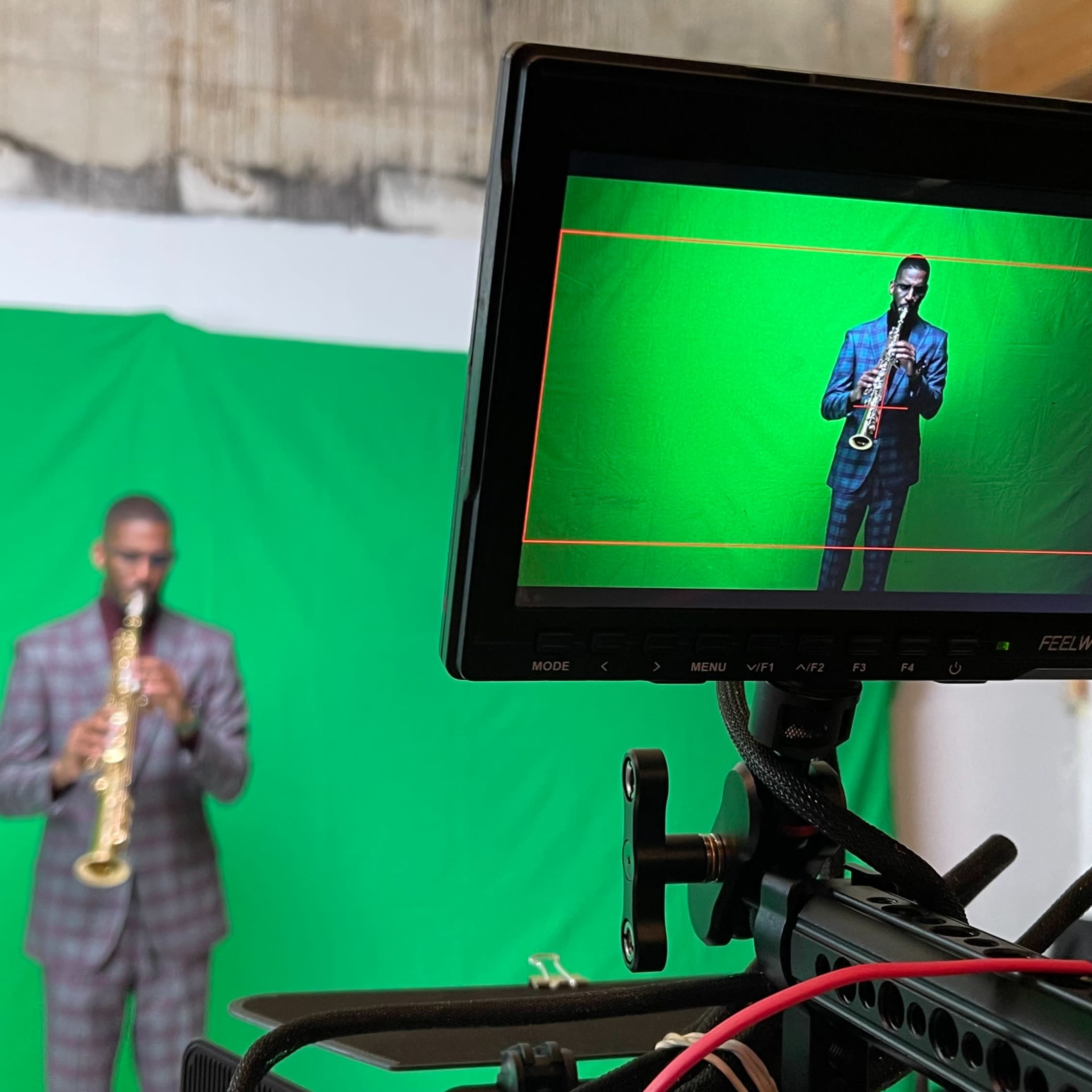 A musician stands in front of a green screen while playing a saxophone, as viewed through a camera monitor.