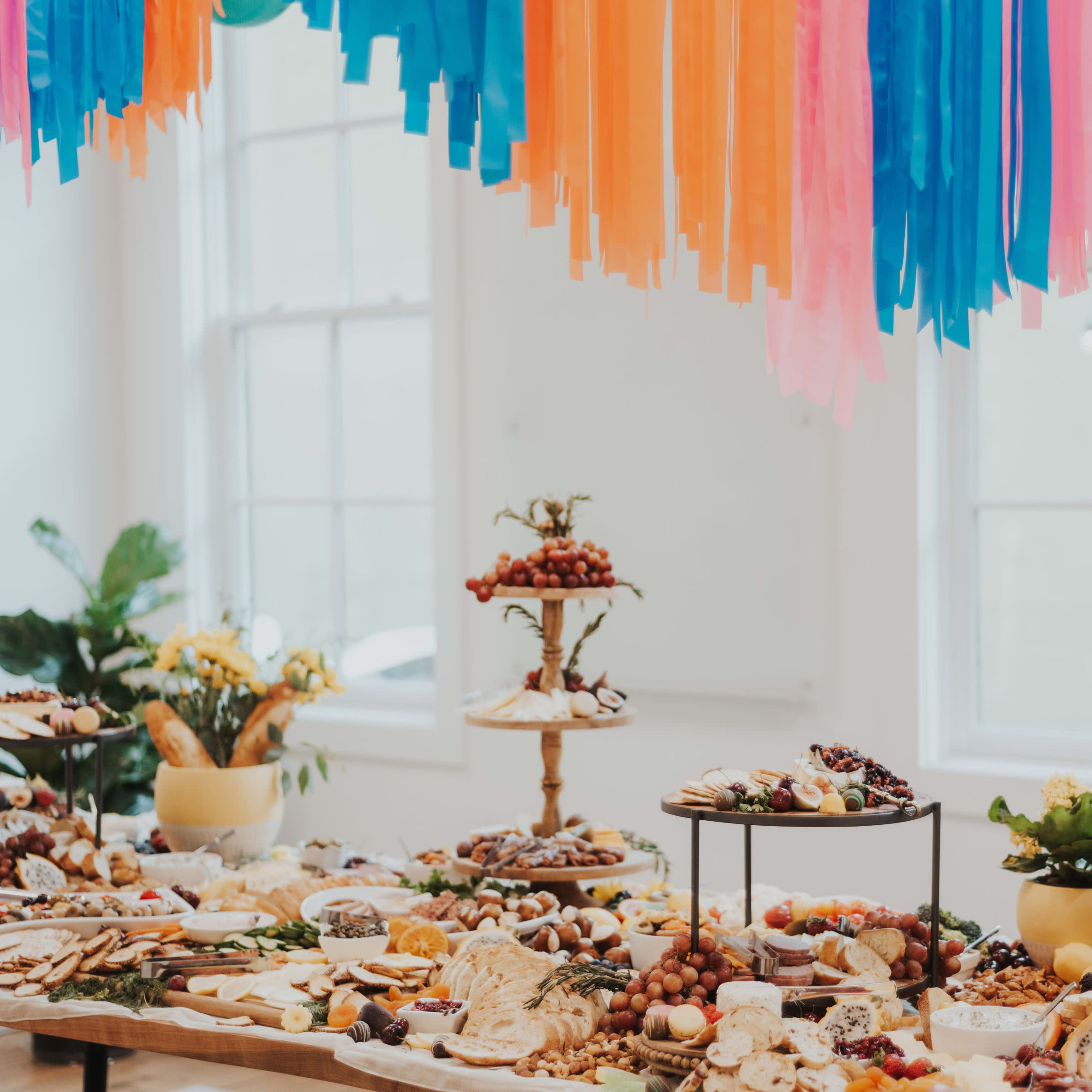 A large spread of assorted foods and snacks is elegantly displayed on tables beneath colorful streamers in a bright room.