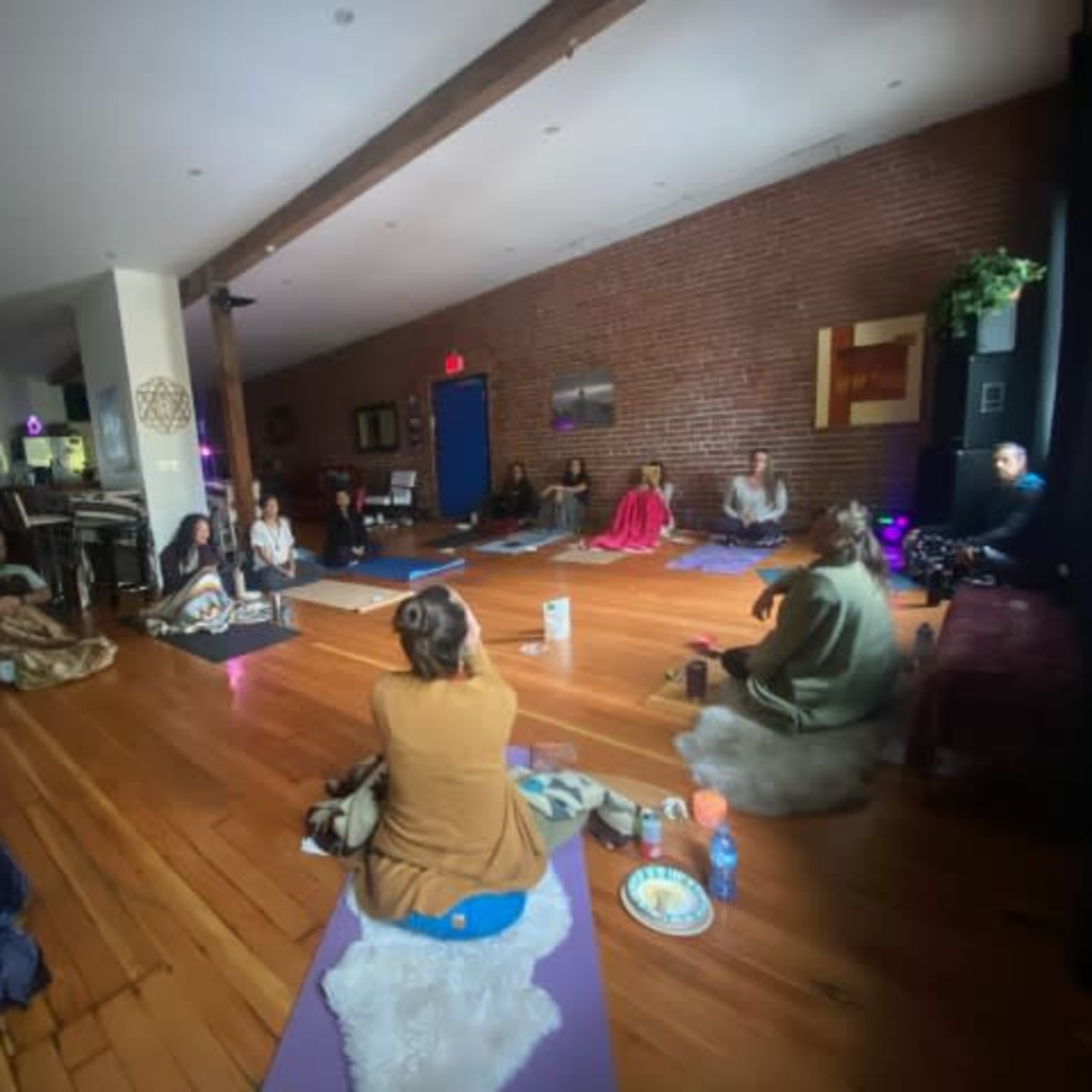 A group of individuals is seated on yoga mats in a spacious room with wooden floors, engaging in a wellness activity.