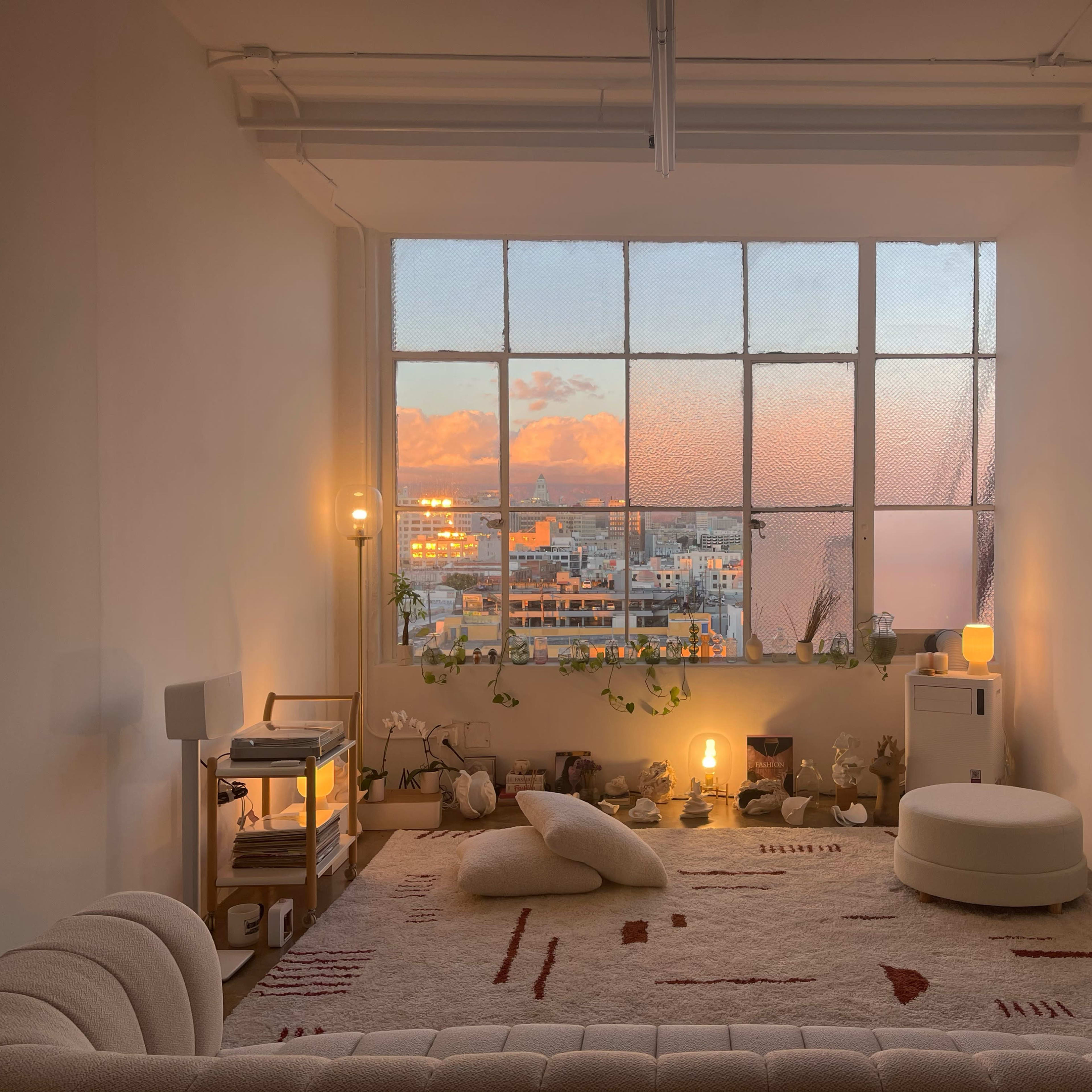 A cozy interior with a large window revealing a sunset view, featuring minimalistic furniture and plants.