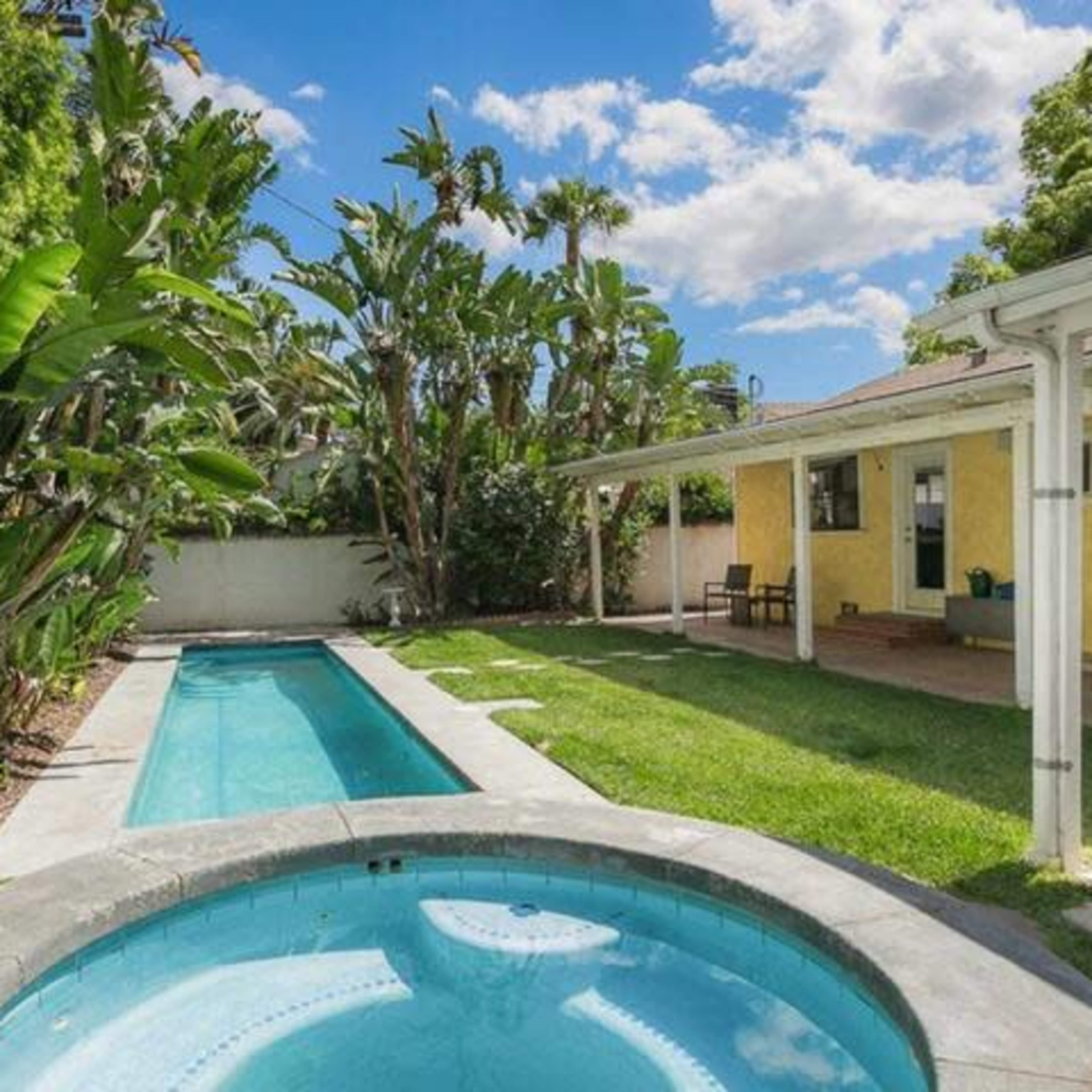 A backyard with a swimming pool, a spa, and a grassy area surrounded by tropical plants and a house with yellow walls.
