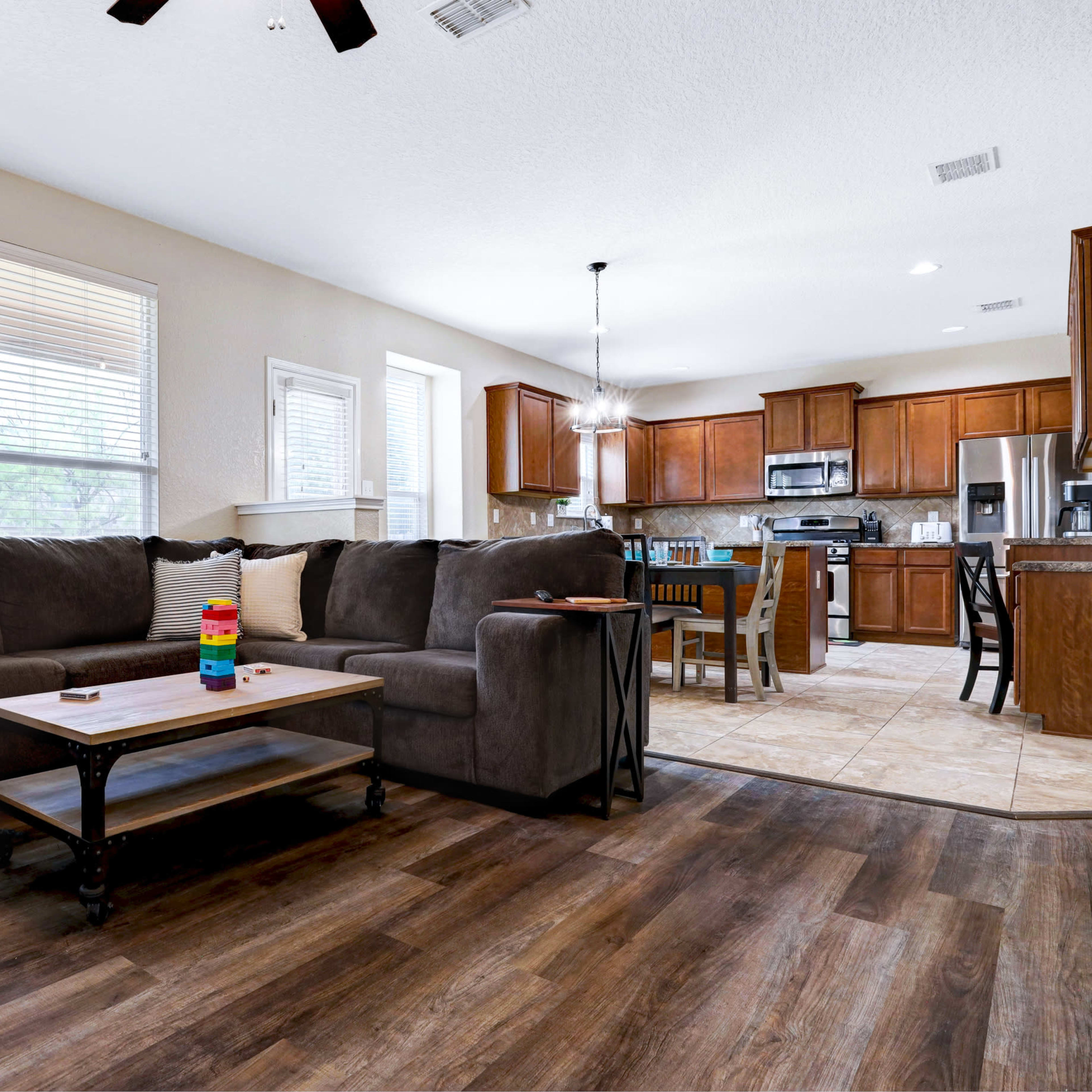 A living room featuring a large gray sectional sofa, a coffee table, and a view into a kitchen with wooden cabinets and stainless steel appliances.