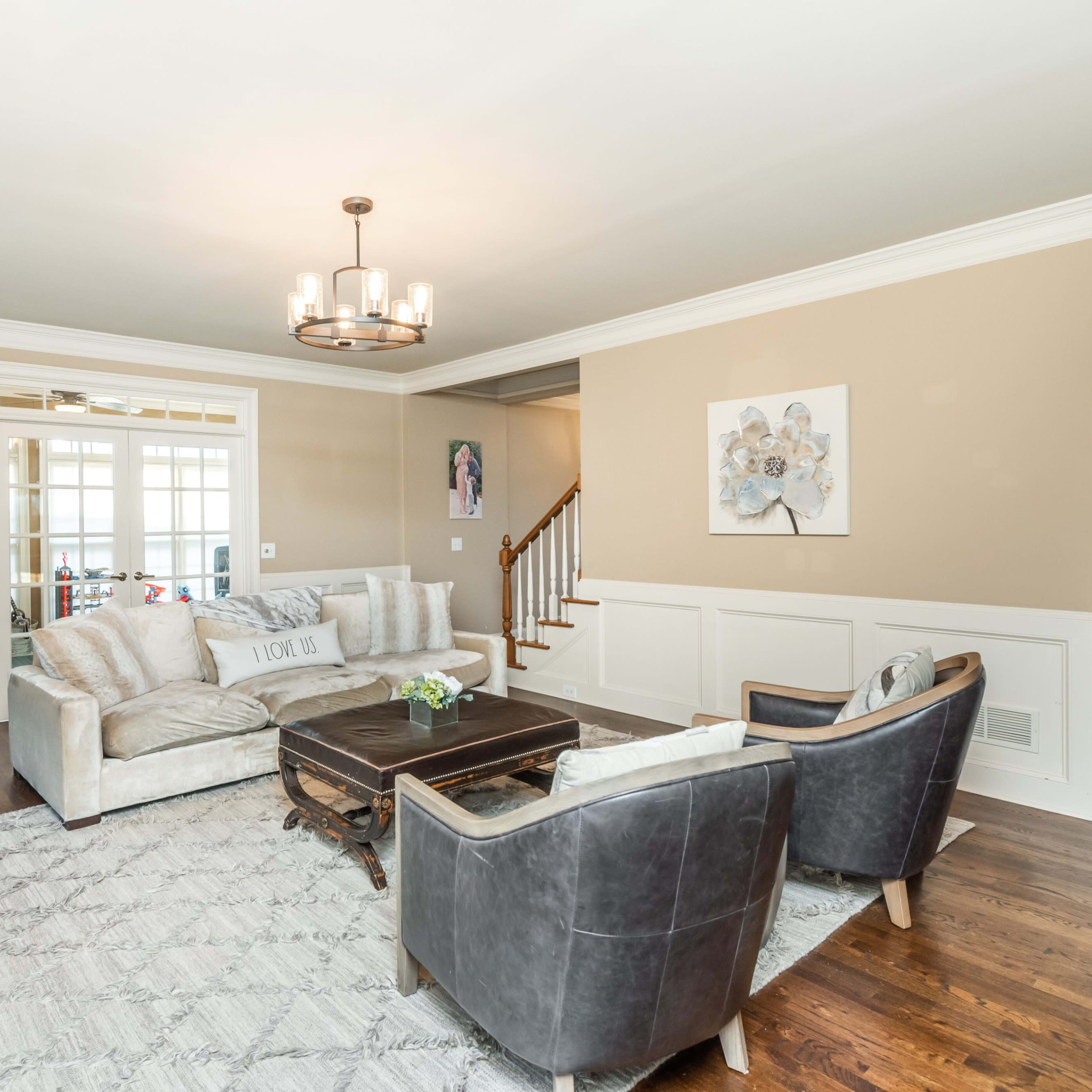 A light-filled living room features a beige sofa, dark chairs, a wooden coffee table, and a decorative wall with wainscoting.
