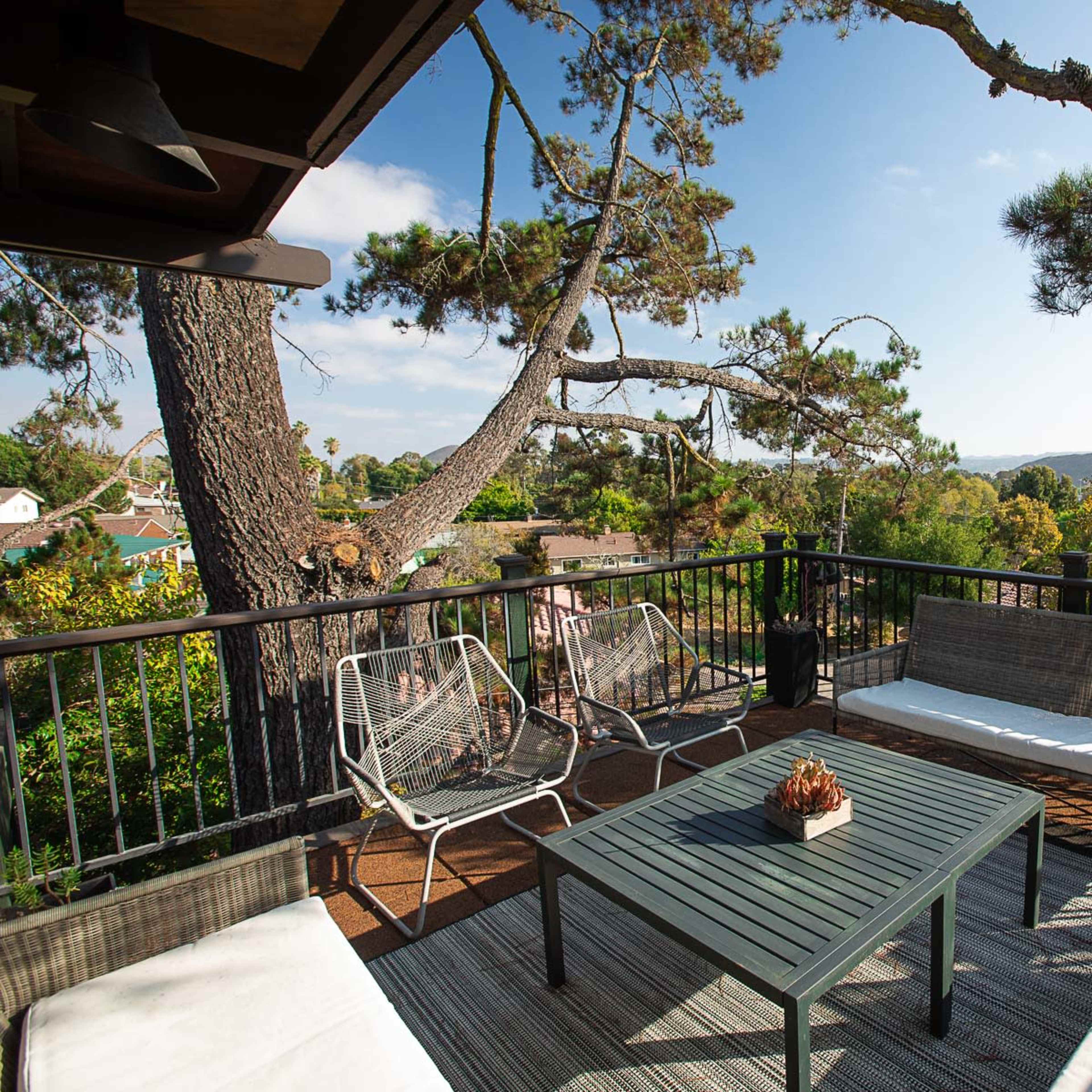 A cozy outdoor seating area features a table and several chairs on a wooden deck surrounded by trees and a distant view of hills.