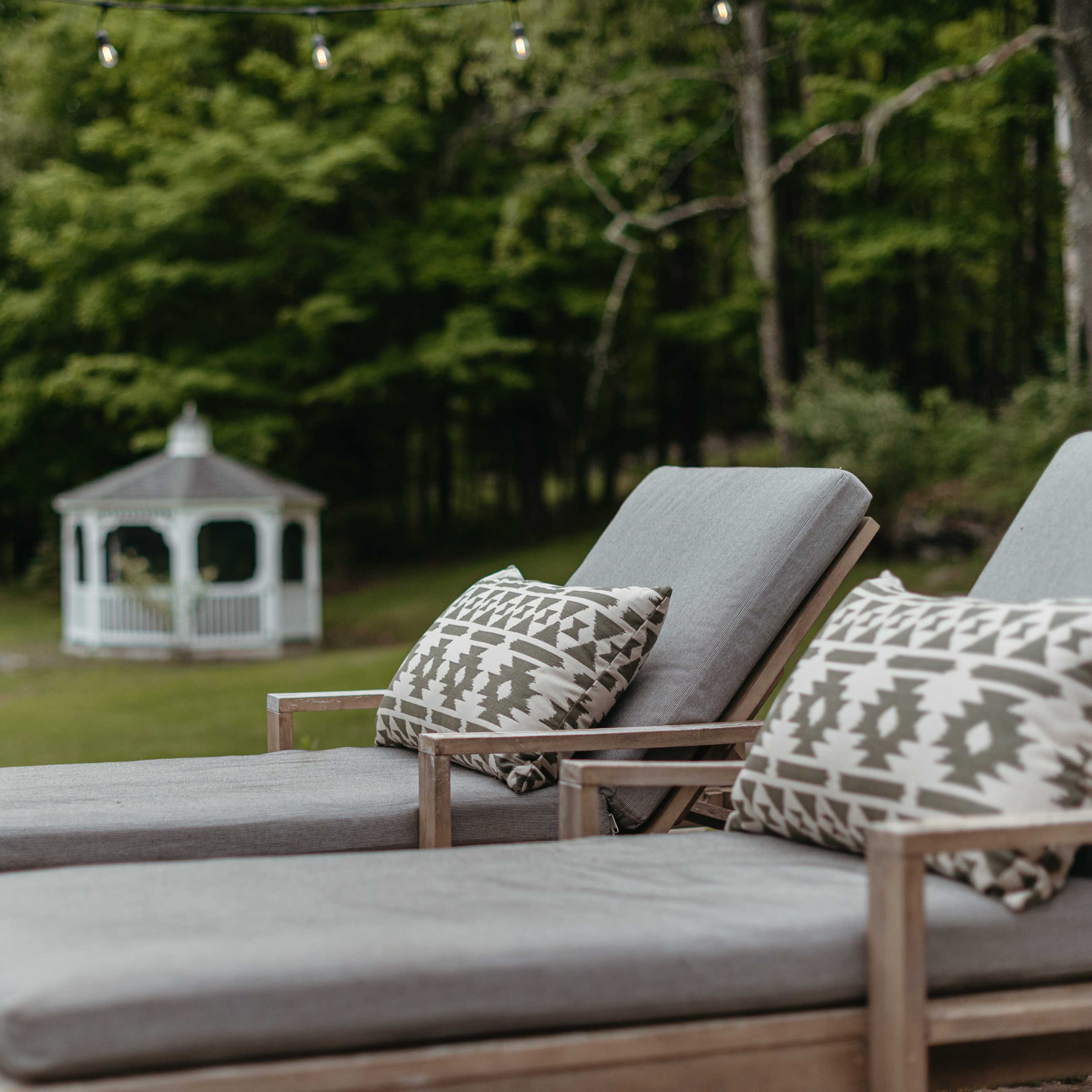 Two wooden lounge chairs with patterned cushions are positioned near a gazebo in a lush green garden.