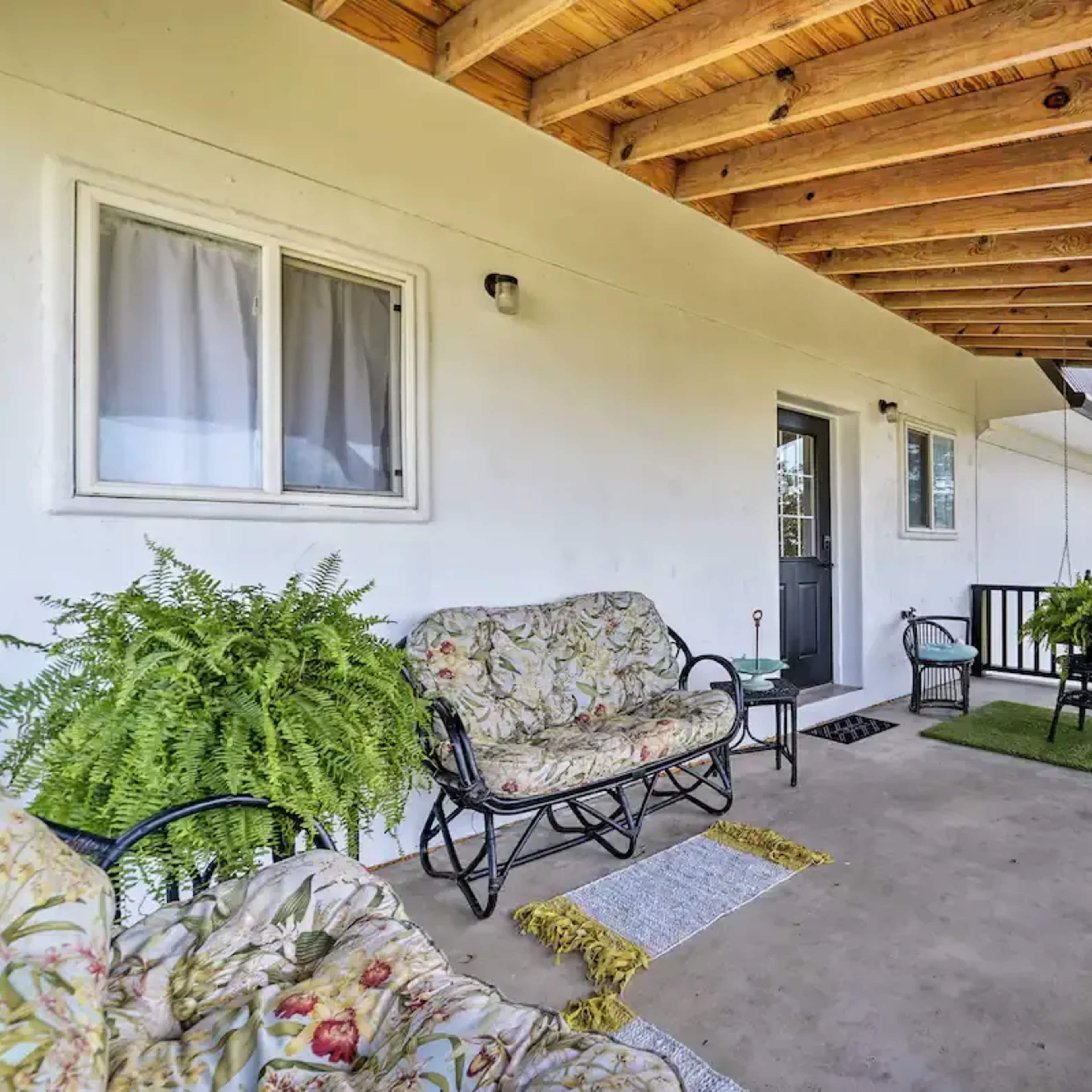 A covered porch with a seating area that includes a floral-patterned sofa and chairs, surrounded by greenery.