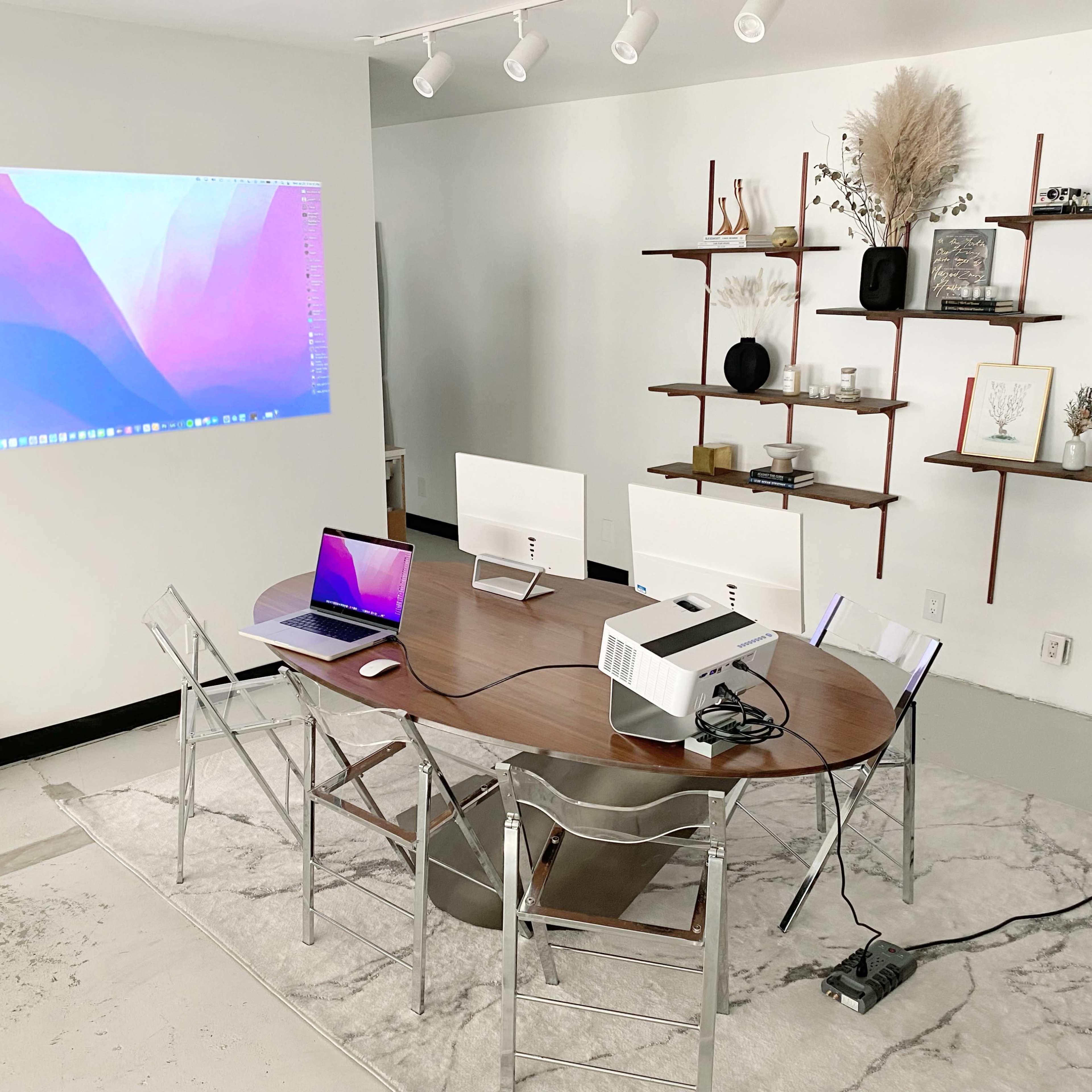 A modern conference room features a wooden oval table with two laptops, a projector, and several shelves displaying decorative items against a light-colored wall.