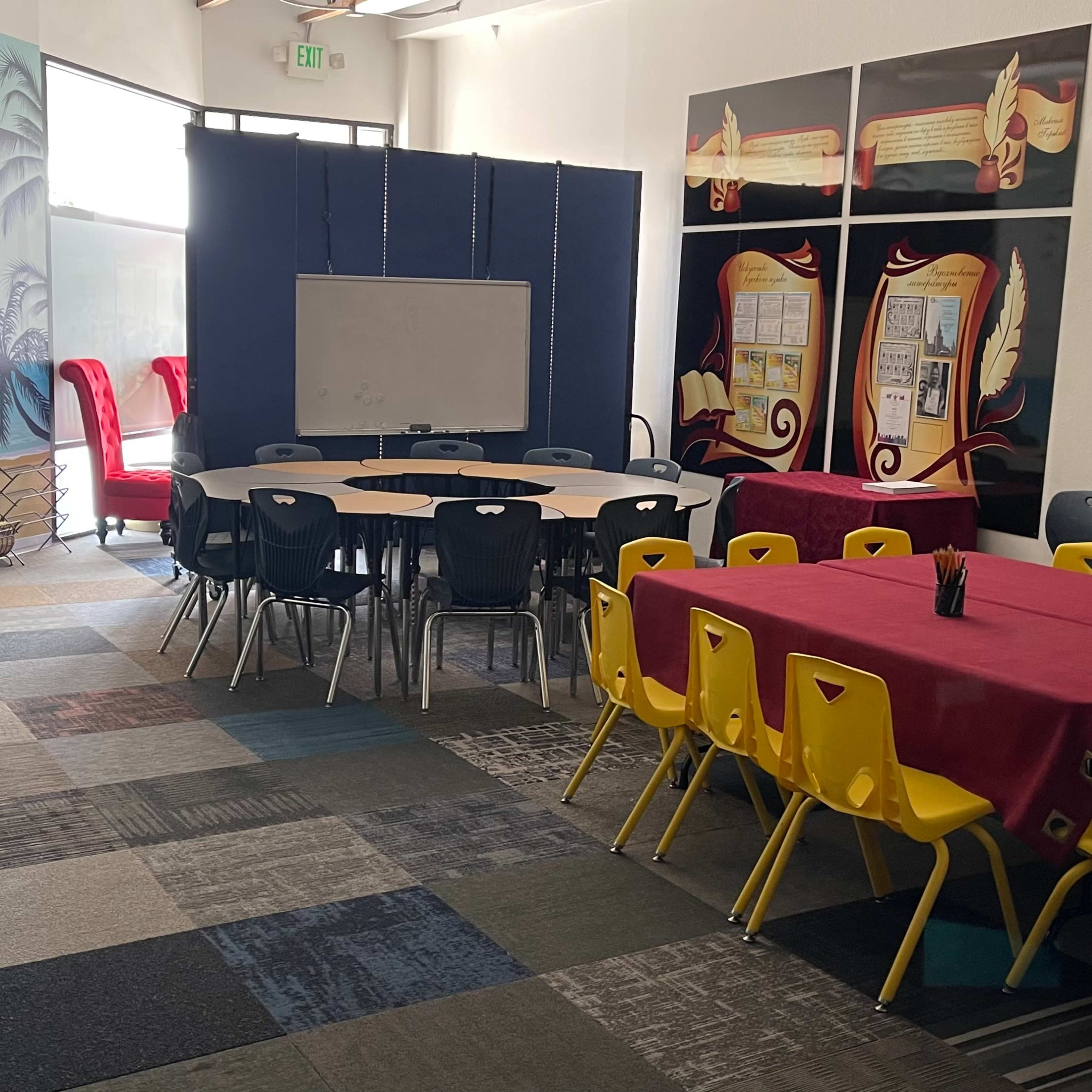 A brightly colored room with several round tables surrounded by yellow chairs, a large whiteboard, and a mural depicting a beach scene.