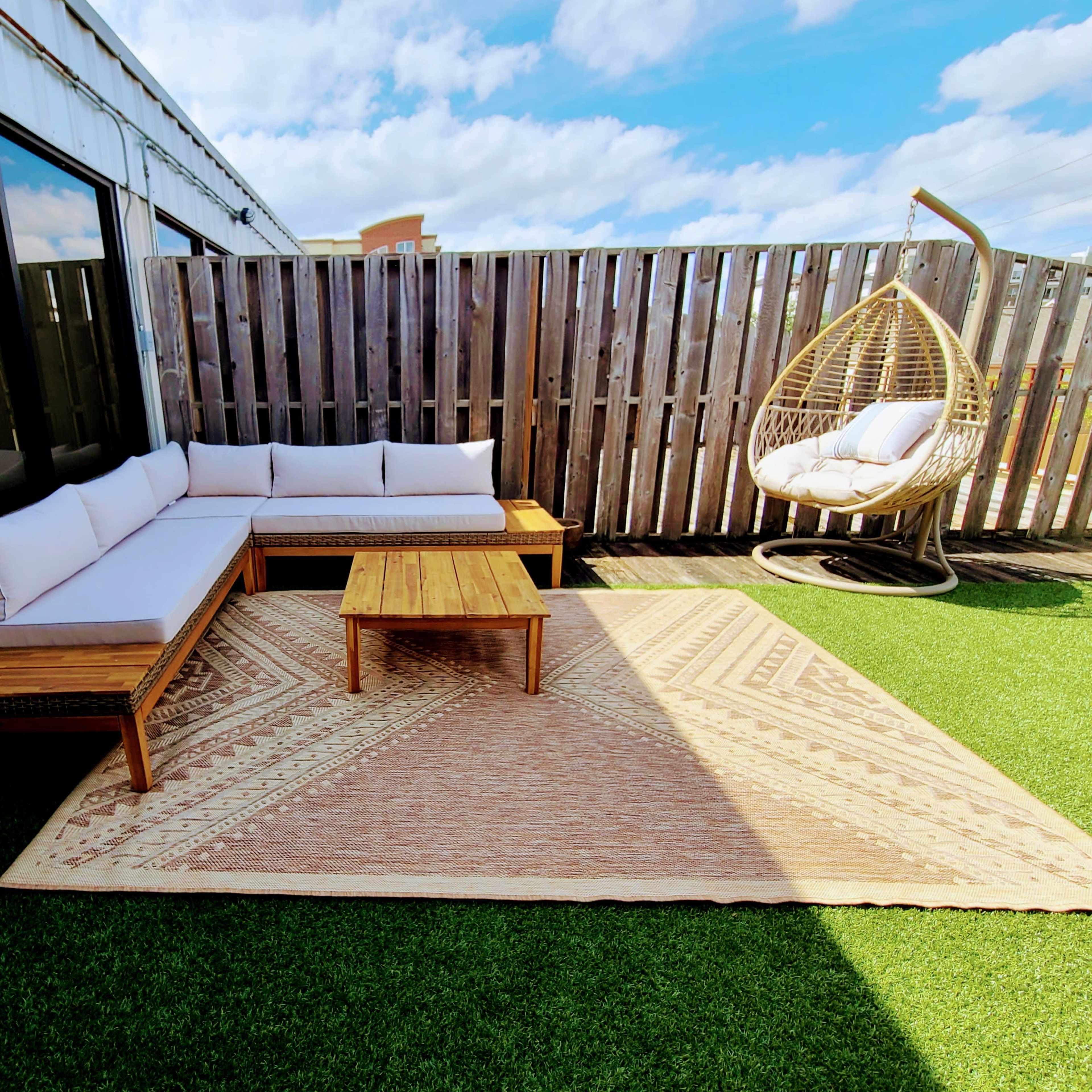 A modern outdoor seating area with a sectional sofa, a wooden coffee table, a hanging chair, and a patterned rug on artificial grass.