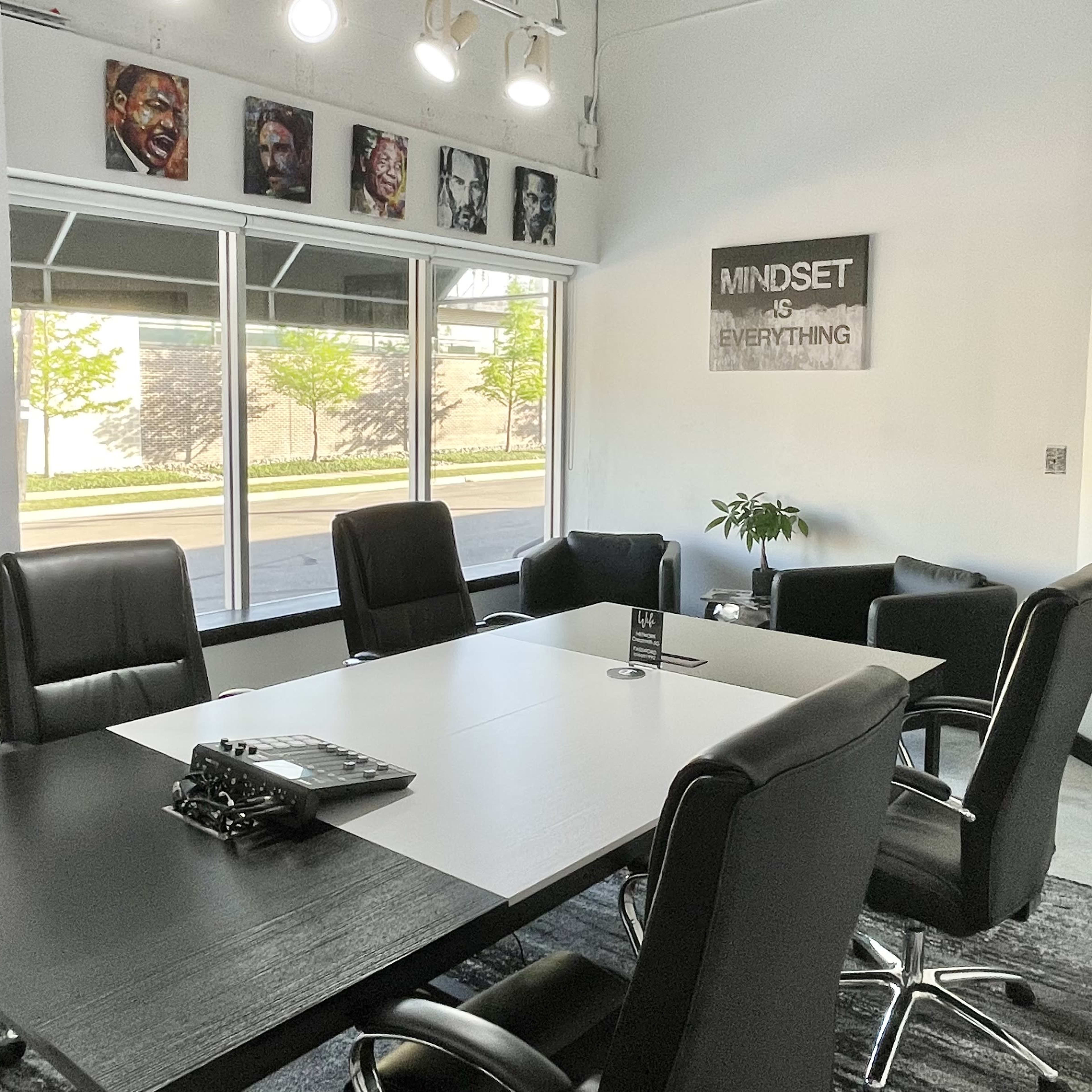 A modern conference room with a large table surrounded by black chairs, featuring windows that provide natural light and a poster on the wall that reads "MINDSET IS EVERYTHING."