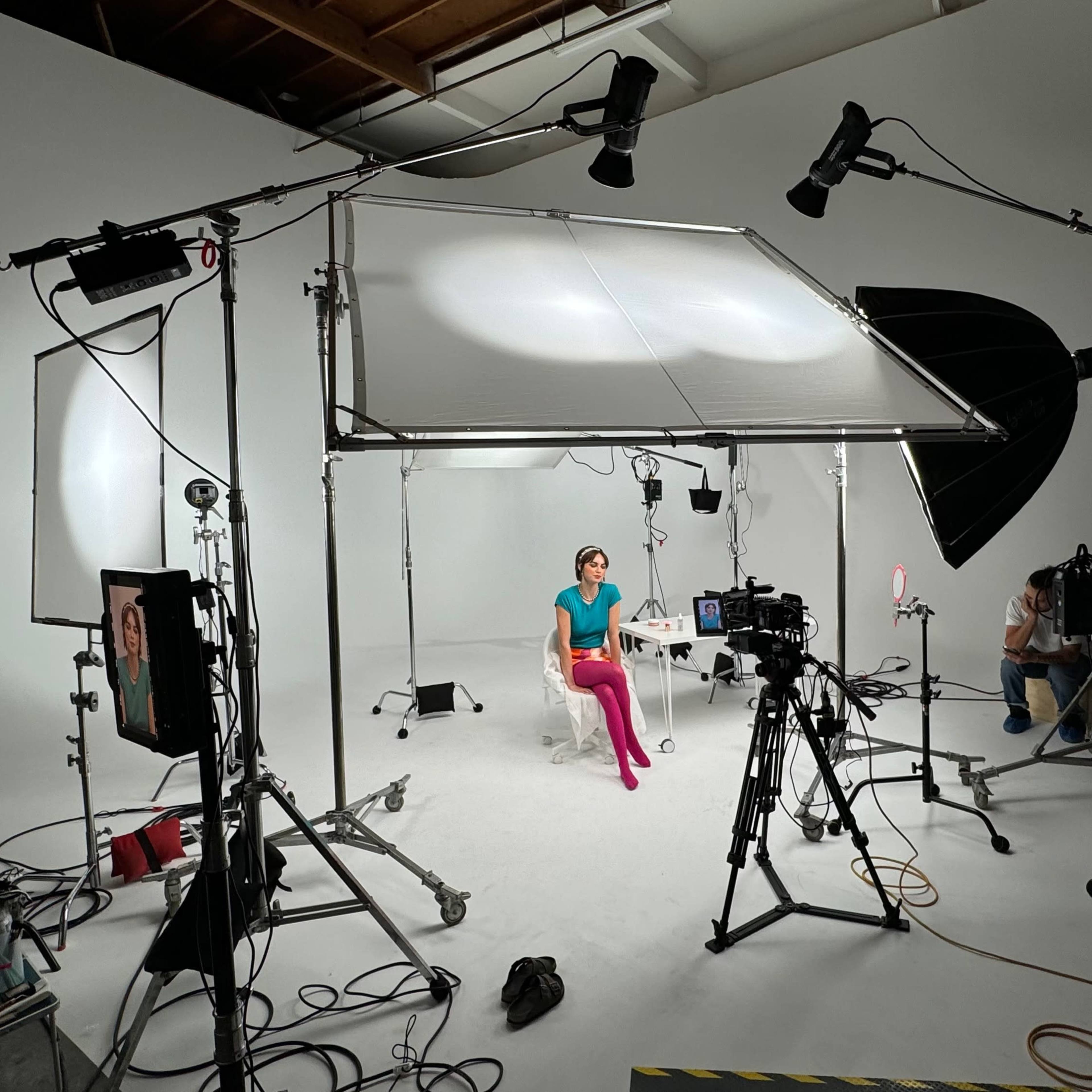 A studio setup features a seated model with bright pink pants, surrounded by various lighting equipment and cameras against a white backdrop.