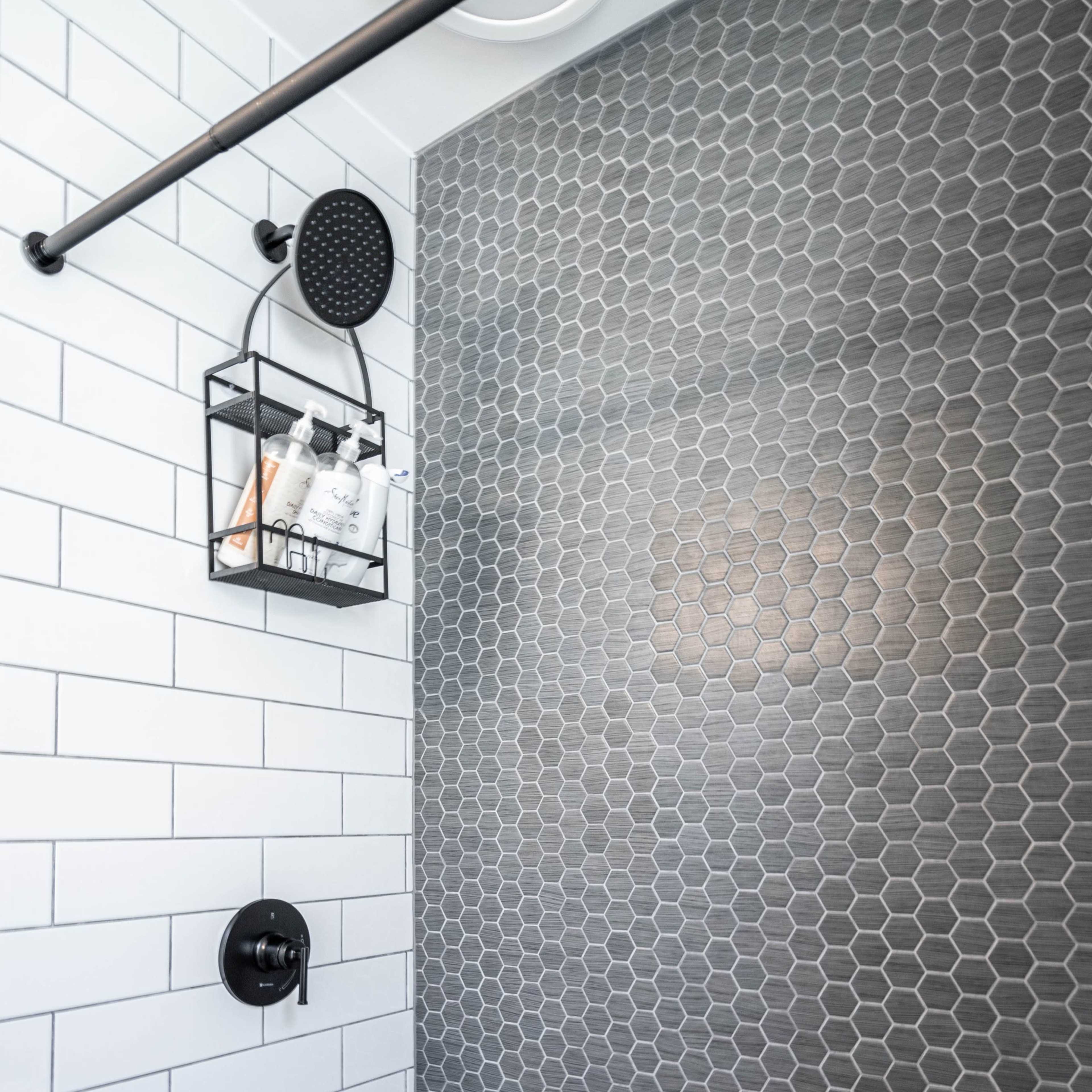 A modern bathroom with a white tiled bathtub and a wall featuring gray hexagonal tiles, along with a black showerhead and a wall-mounted storage basket.