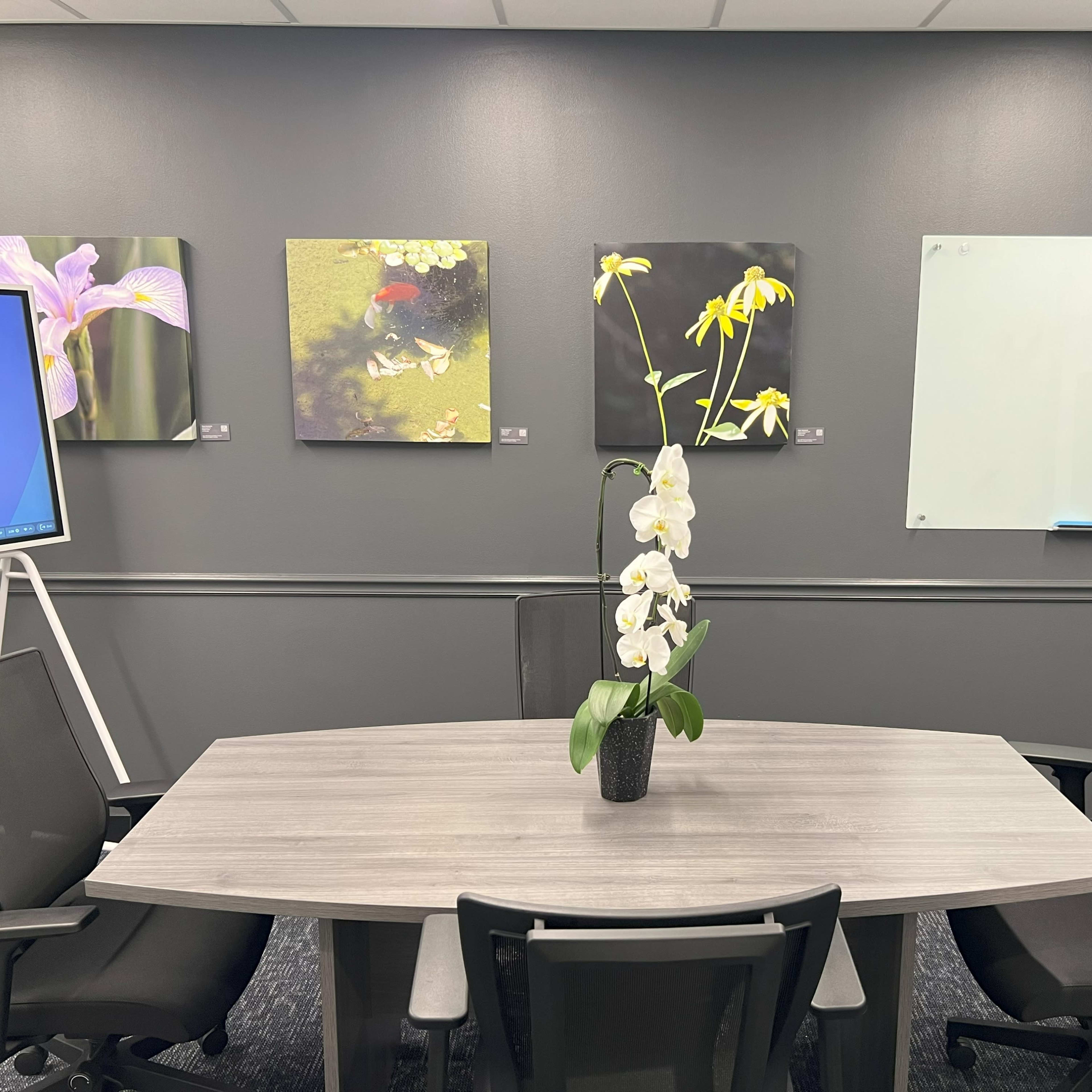 A conference room features a round wooden table surrounded by black chairs, with floral artwork displayed on the gray walls and a whiteboard in the corner.