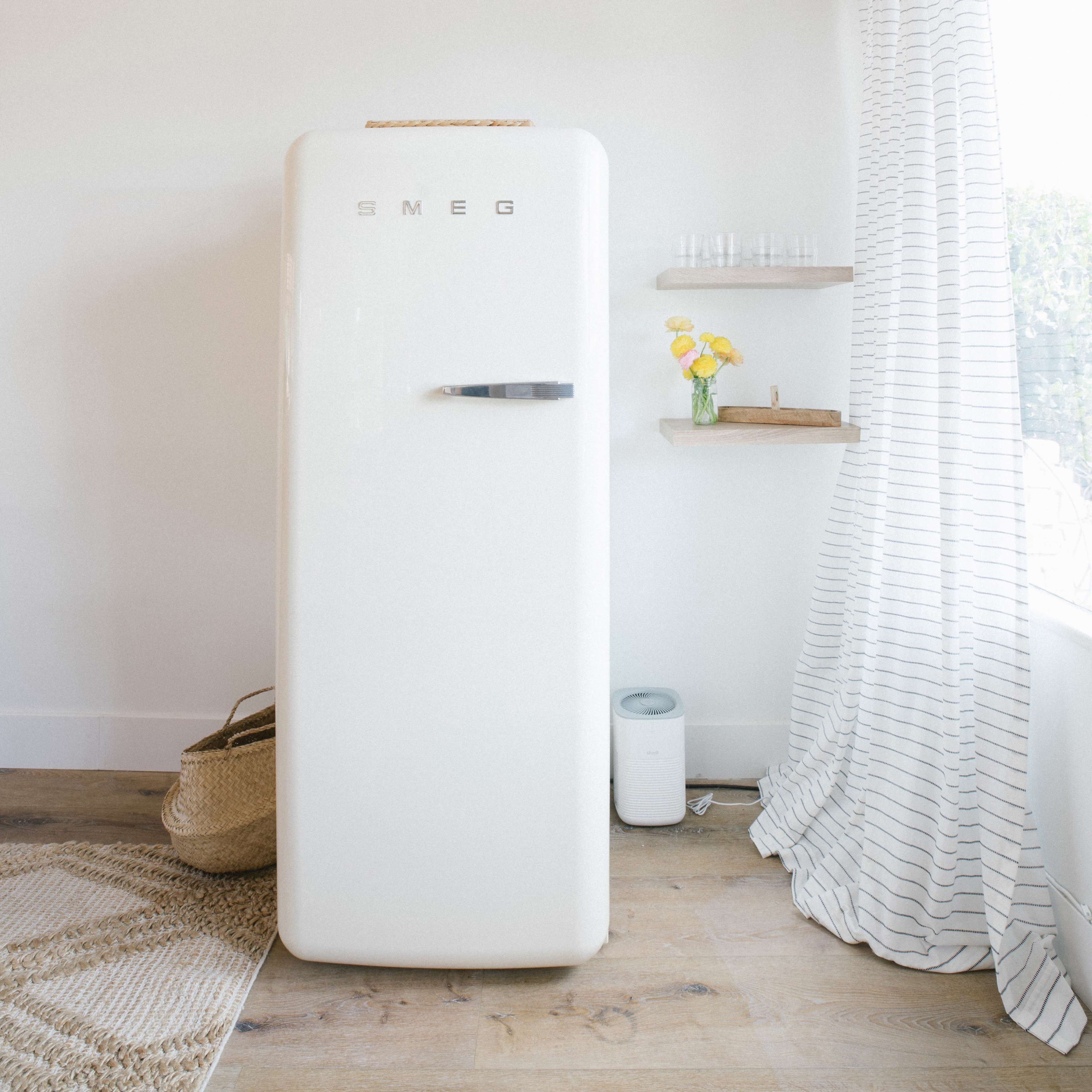 A white retro-style fridge stands against a wall next to a decorative shelf with a small vase of flowers and a woven basket on the floor.