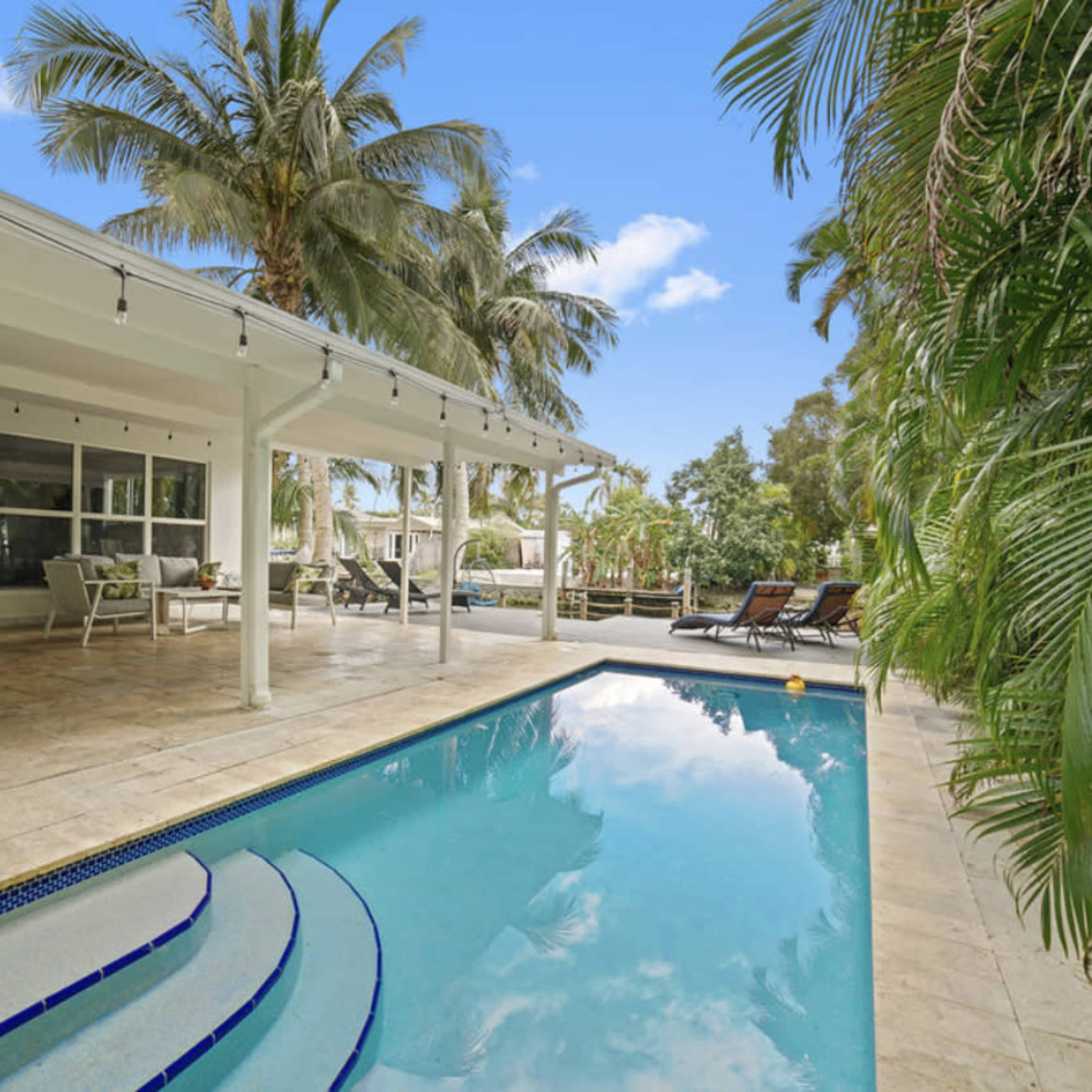 A landscaped backyard with a swimming pool and lounging area, surrounded by palm trees and outdoor seating.