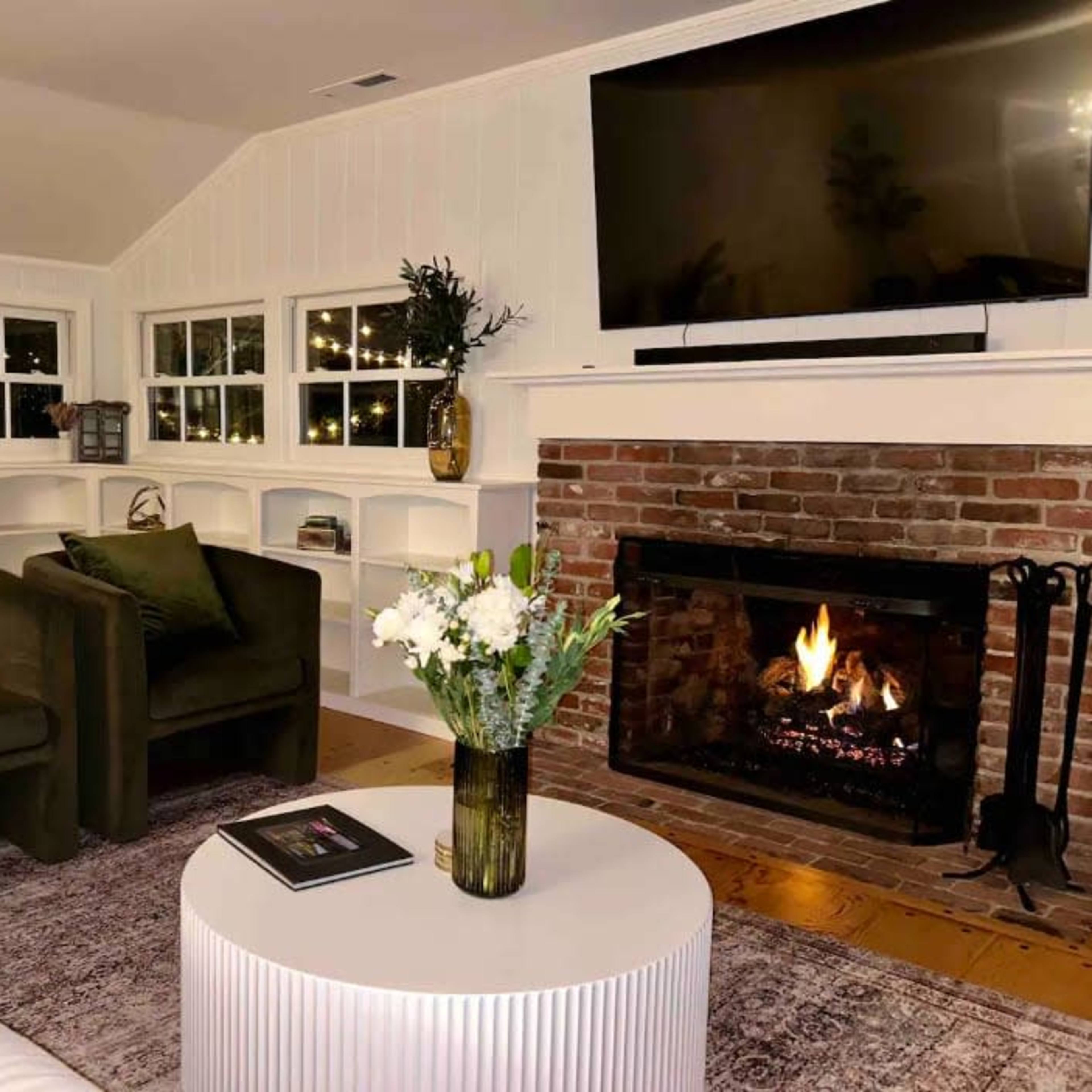 A cozy living room features two dark green chairs, a round white coffee table, and a brick fireplace with a lit fire, surrounded by built-in shelves and windows.
