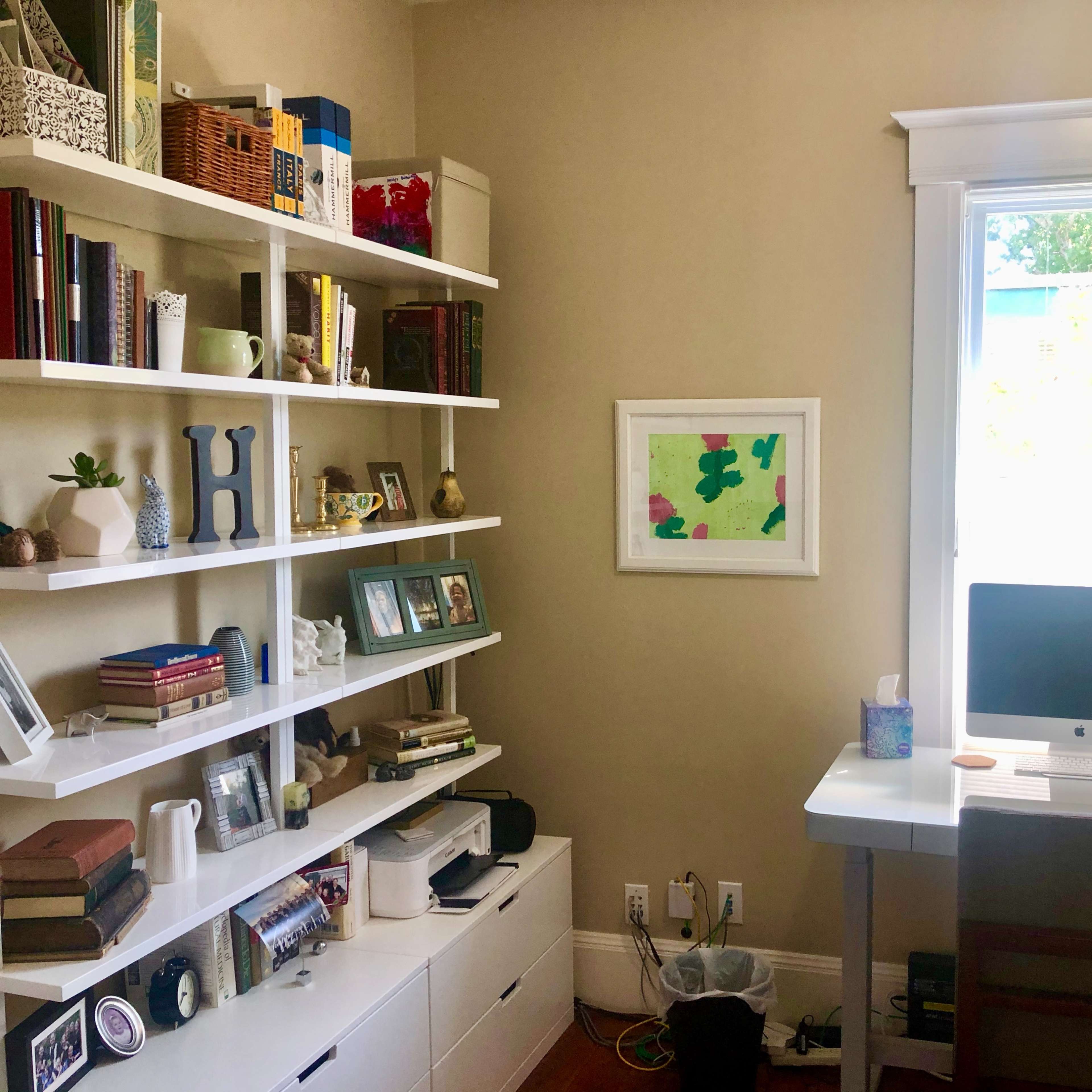 A cozy home office with a white desk, an iMac computer, and a bookcase filled with books and decorative items, beside a window that lets in natural light.