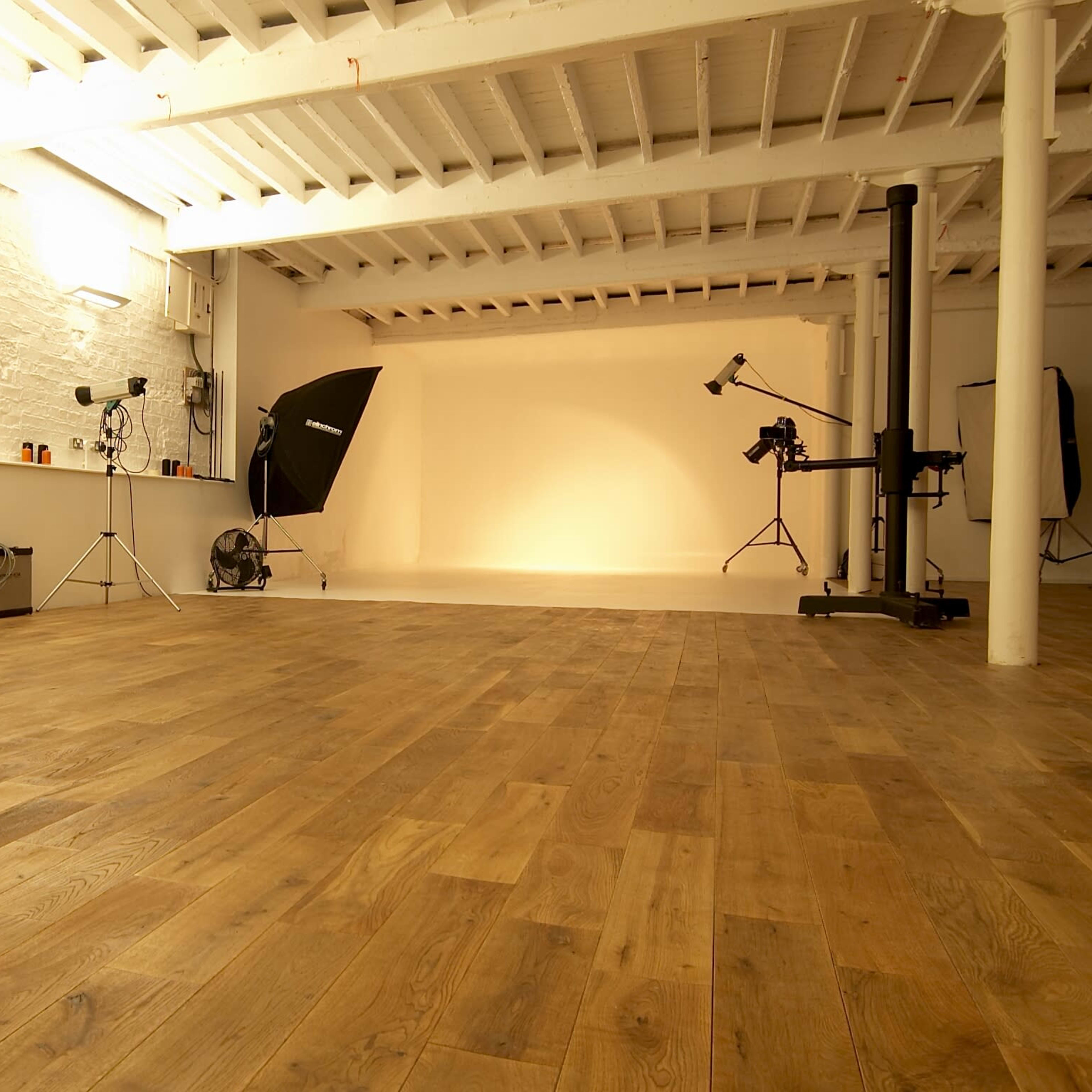A spacious photography studio with wooden flooring, white walls, and various lighting equipment arranged around a neutral backdrop.