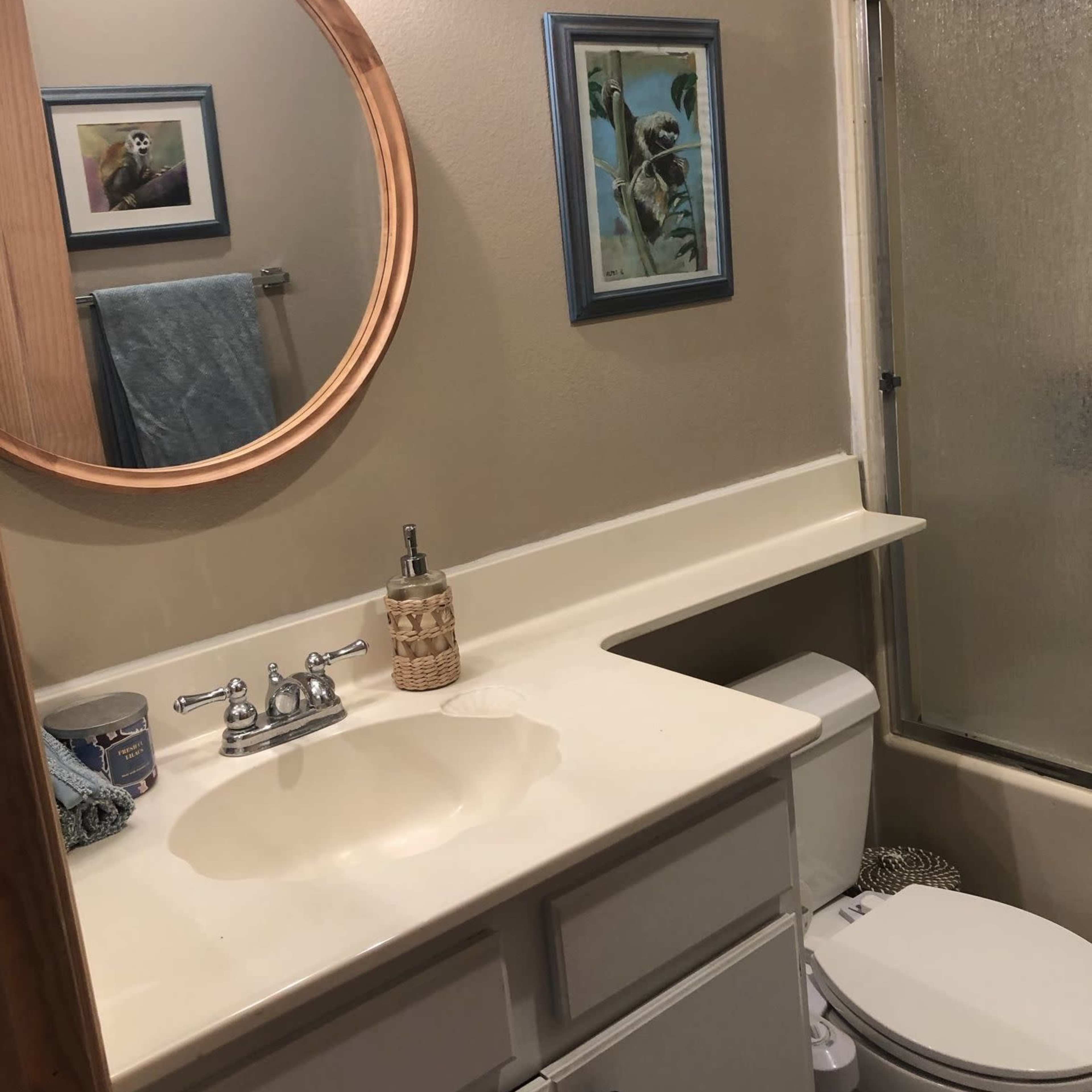 A bathroom with a circular mirror above a sink, a shower area with a glass door, and a framed painting on the wall.