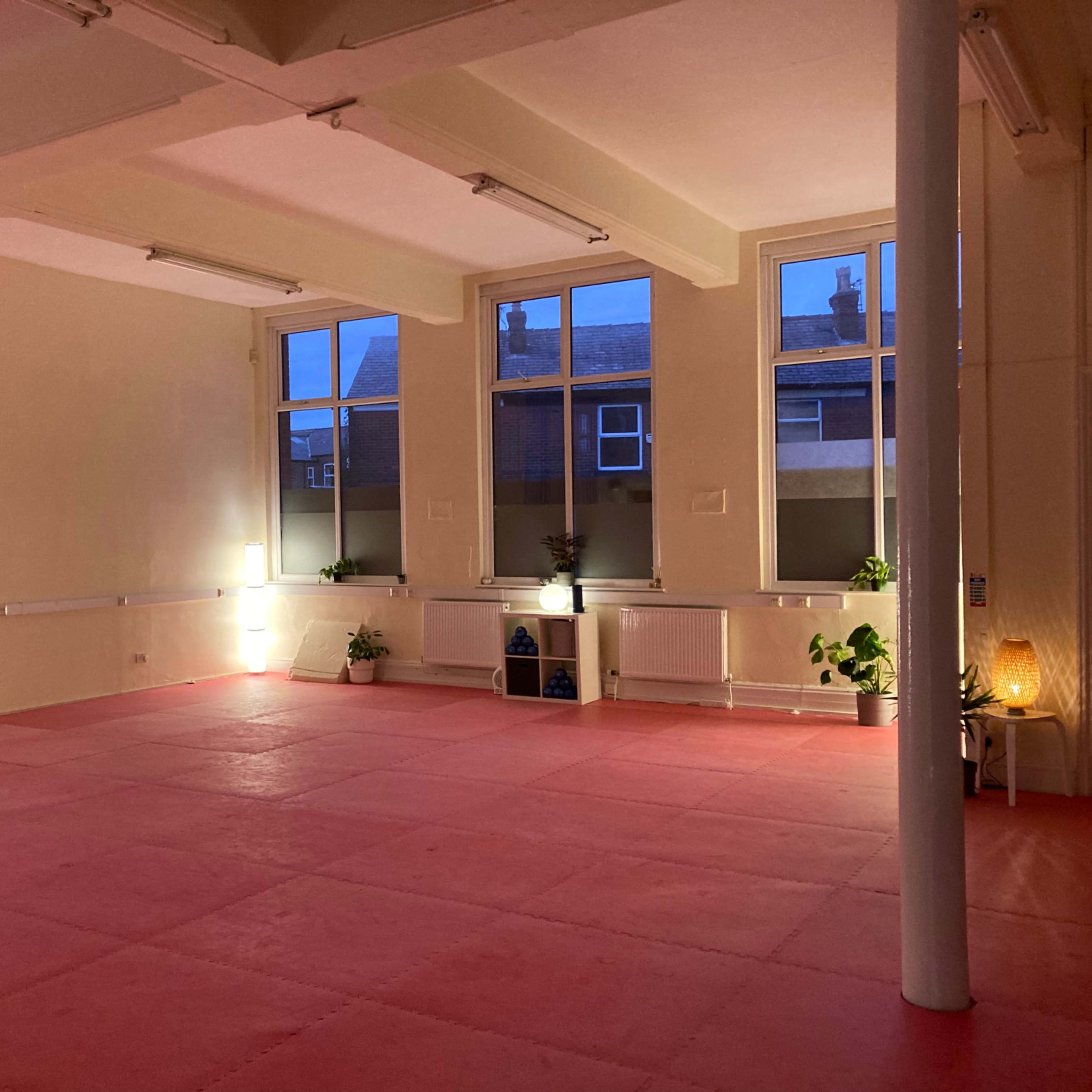 A spacious, empty room with pink flooring, large windows, and minimal lighting, featuring several potted plants.