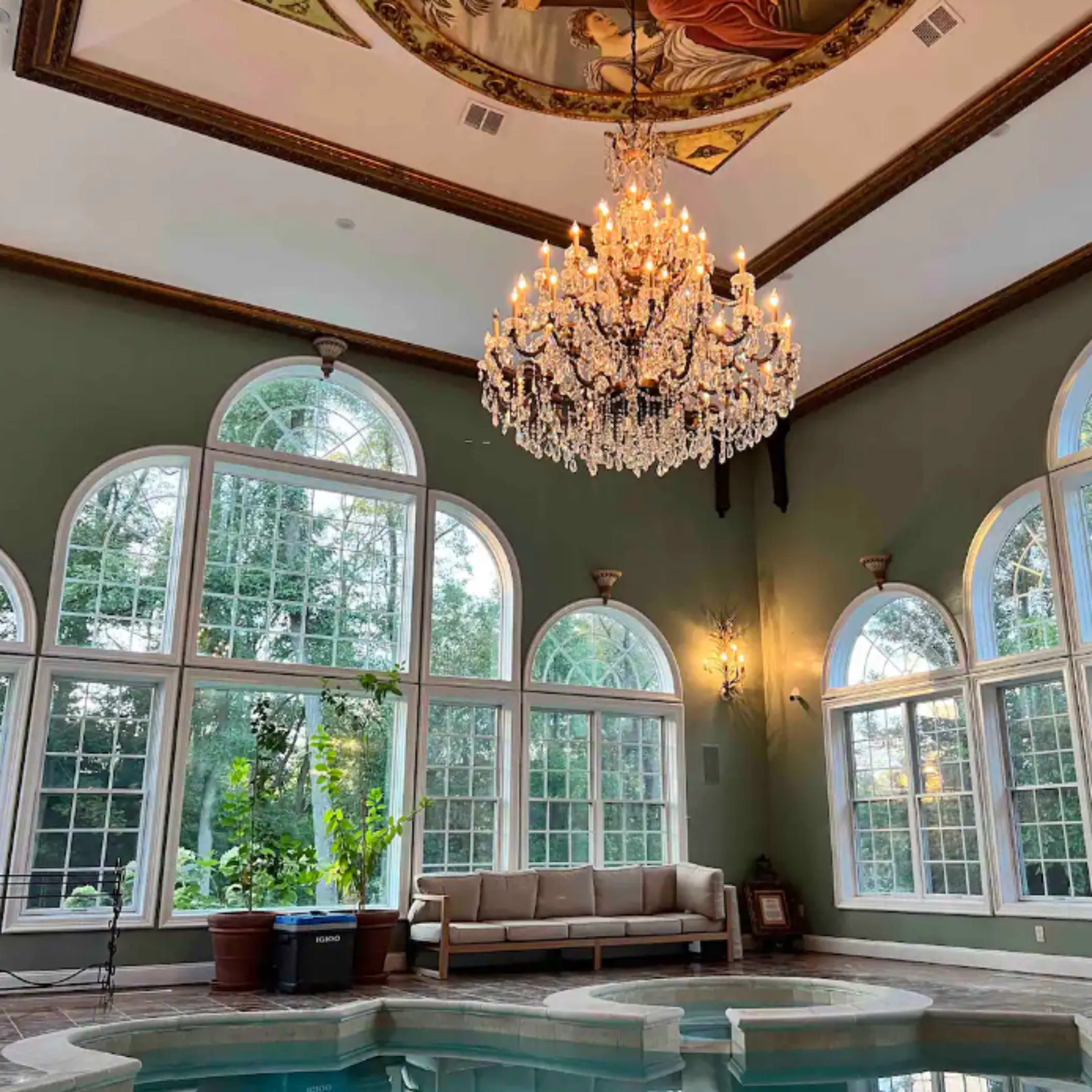 A room with a chandelier, large arched windows, and a small indoor pool surrounded by green walls and a seating area.