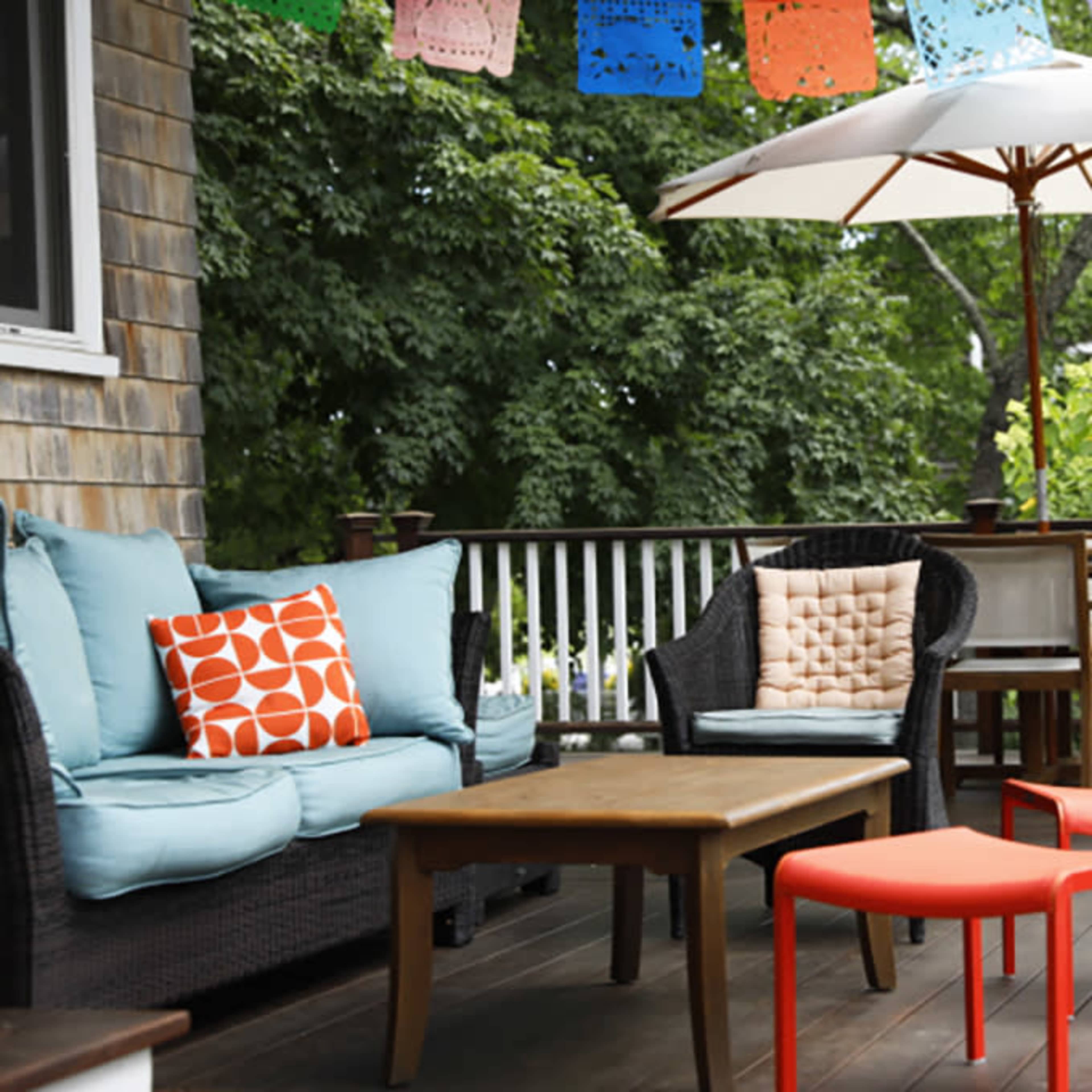 A patio area features a blue cushioned sofa with orange and cream decorative pillows, a wooden table, two red stools, and an umbrella, surrounded by greenery and colorful paper decorations.