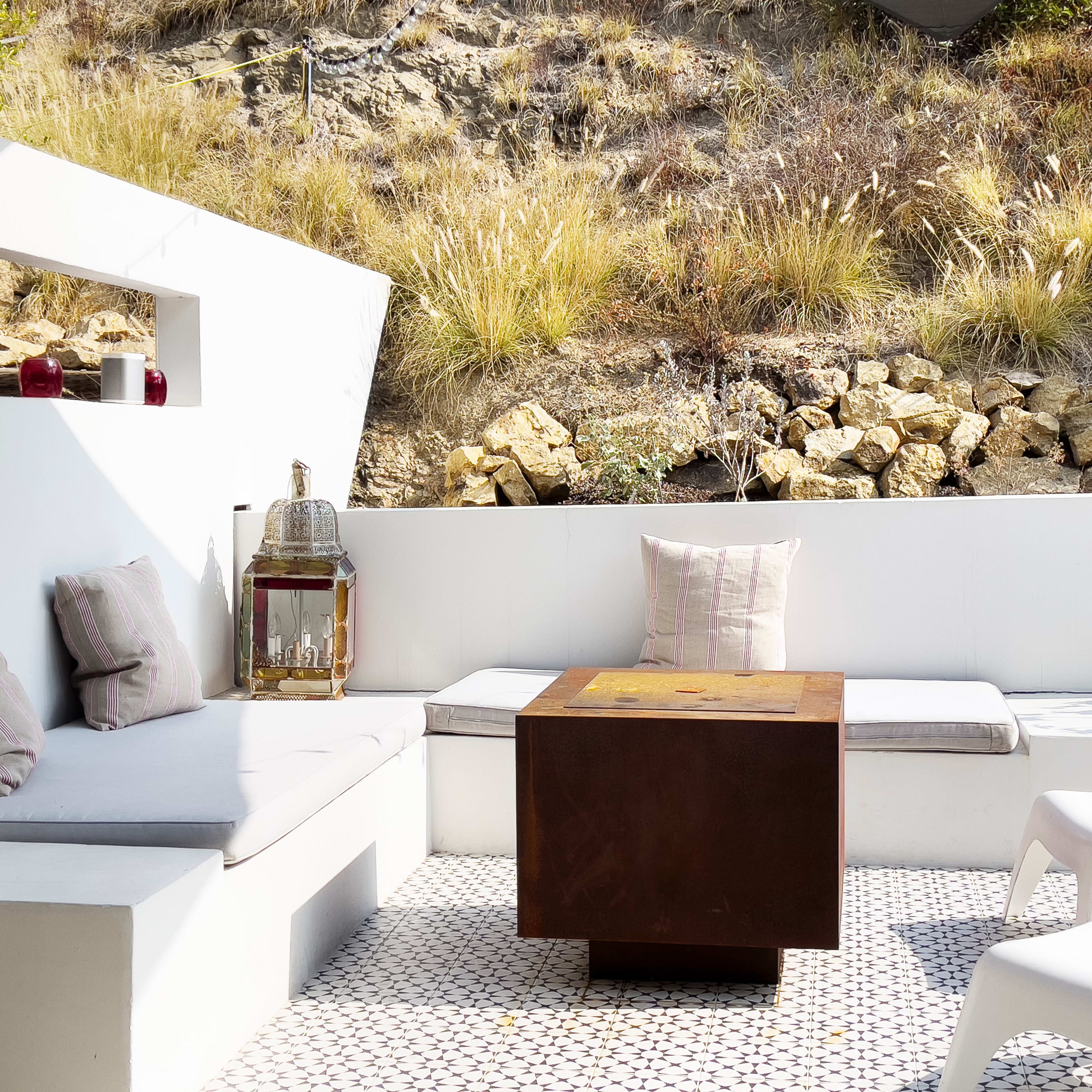 A modern outdoor seating area features a metal coffee table, surrounded by white couches and accented by decorative pillows, set against a backdrop of rocky terrain and dry grasses.