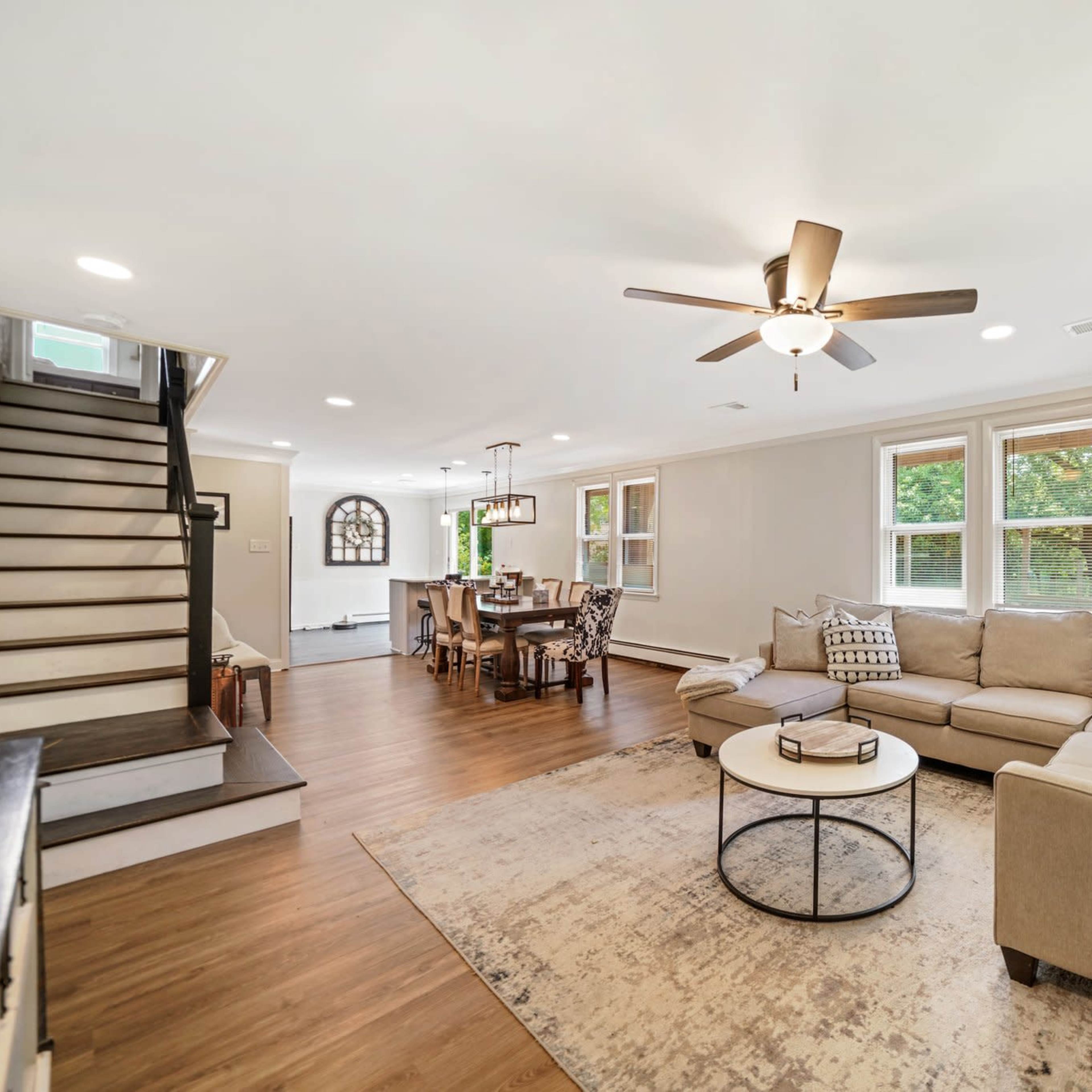 A spacious living room with a sectional sofa, a round coffee table, a ceiling fan, and a staircase leading to an upper level, with dining space visible in the background.