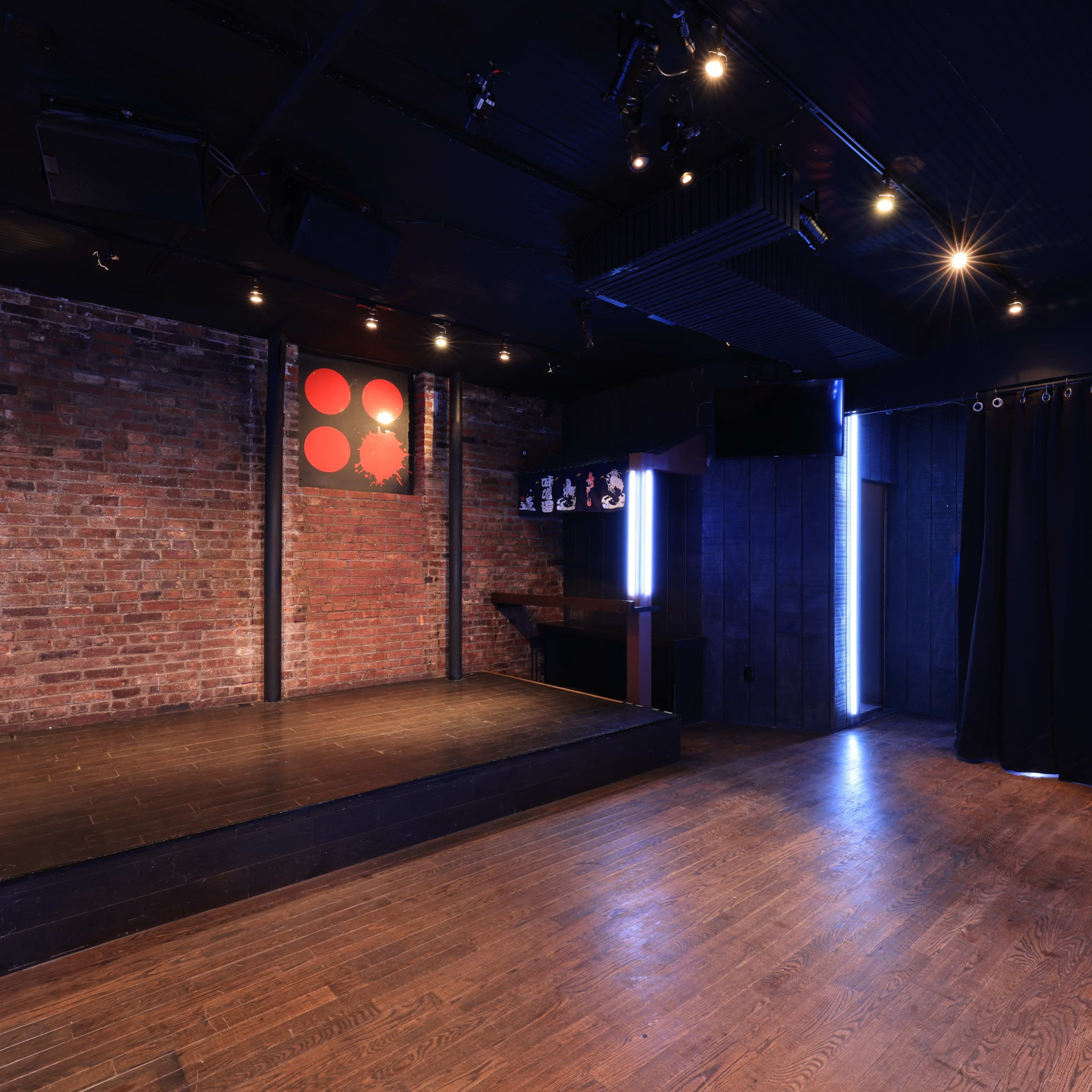 A dimly lit performance space with a wooden stage, exposed brick walls, and red lanterns hanging from the ceiling.