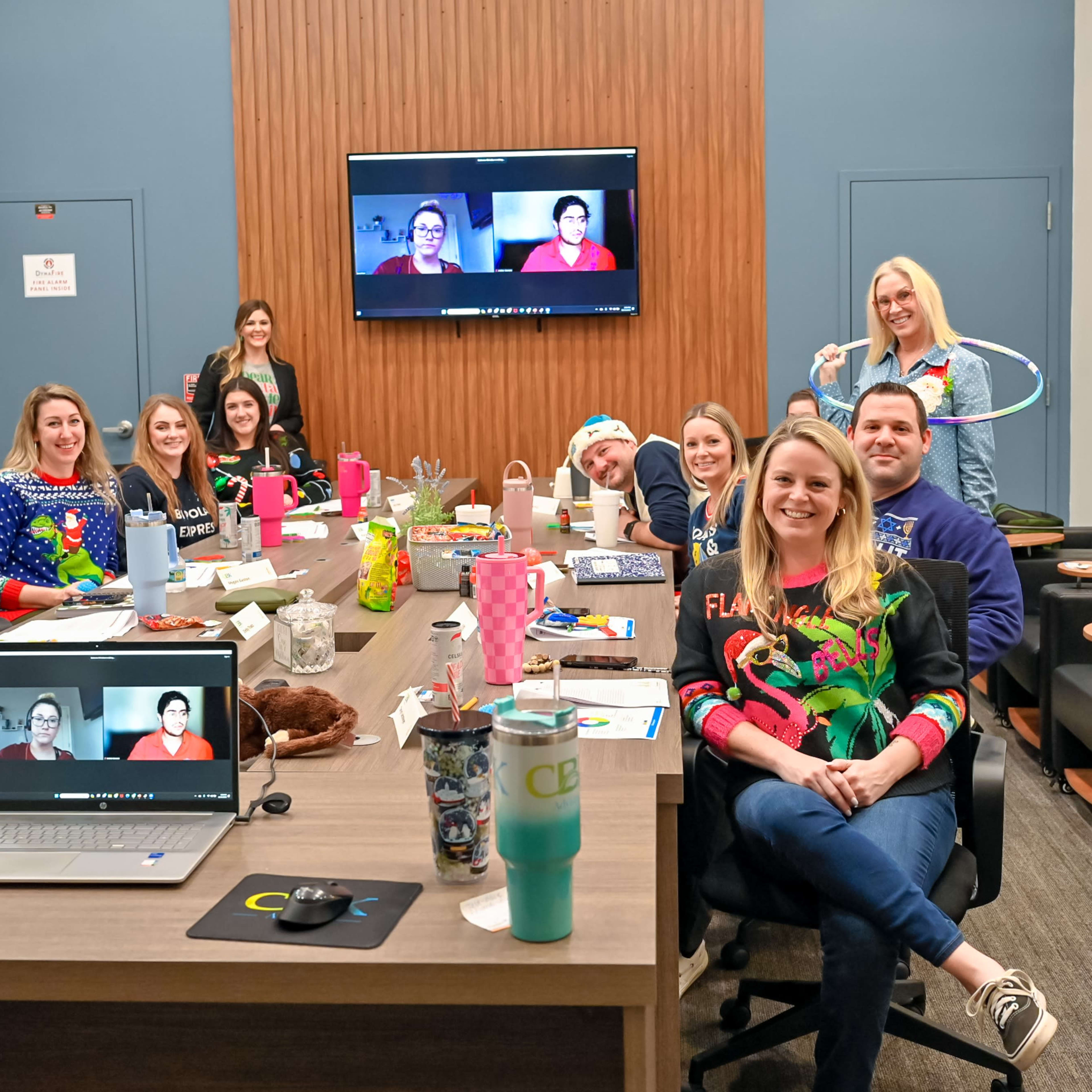 A group of twelve people is gathered around a conference table in a well-lit meeting room, some wearing festive sweaters, while others appear on a video call displayed on a large screen.