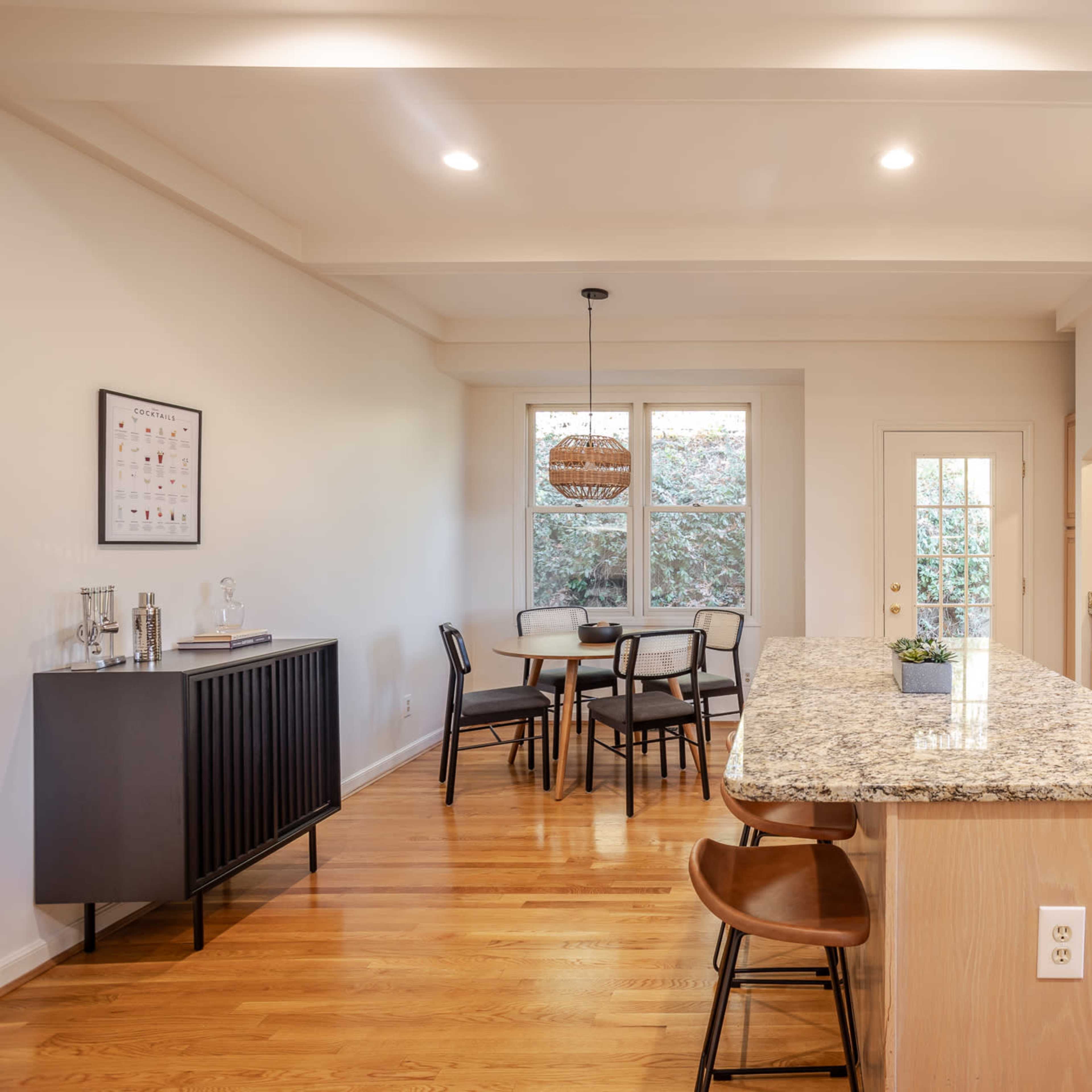 A modern kitchen with wooden cabinets, a granite countertop island, a dining area in the background, and large windows letting in natural light.