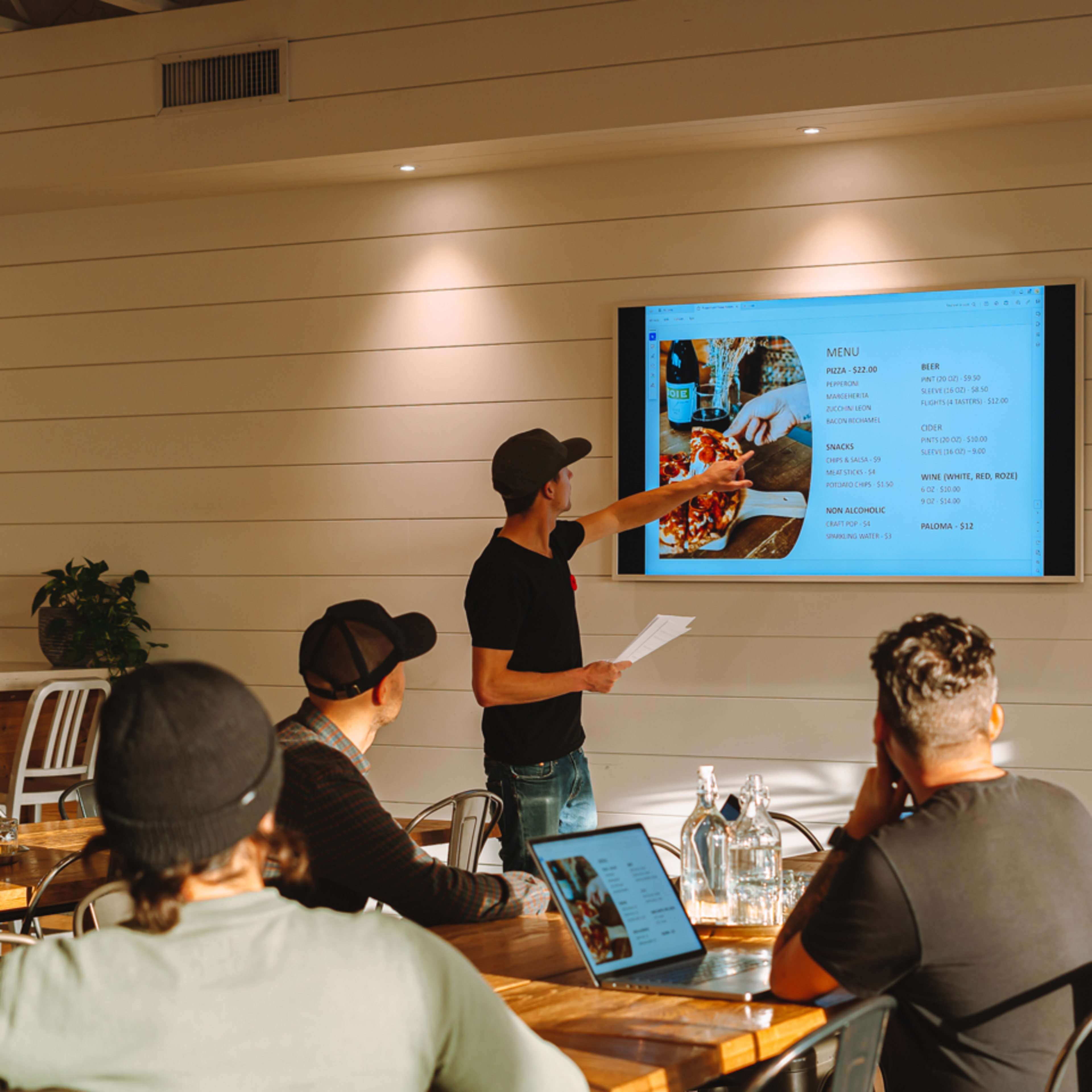 A man points at a presentation screen displaying a menu while three colleagues watch in a bright meeting room.