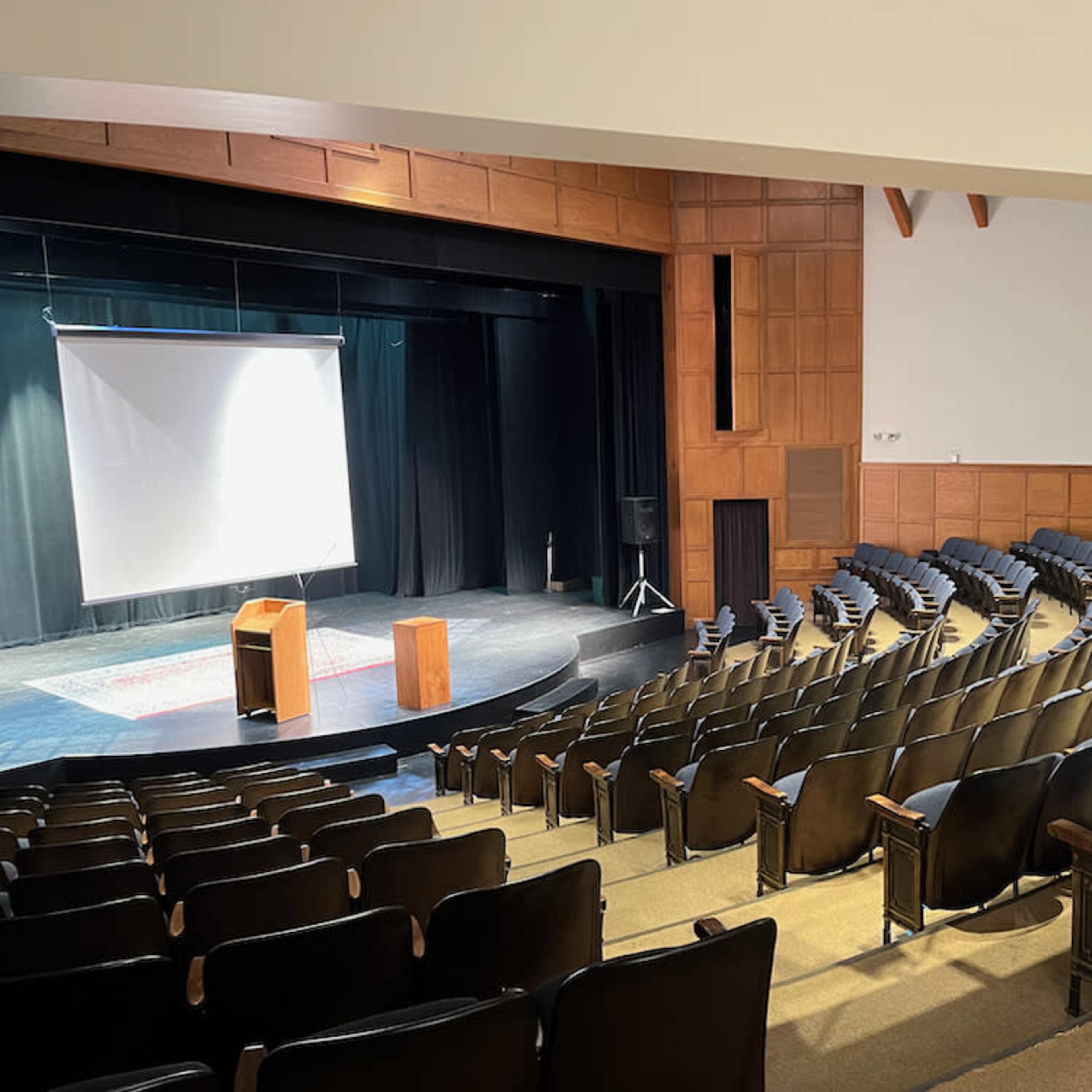 An empty theater with rows of black seats facing a stage equipped with a projection screen and a wooden podium.