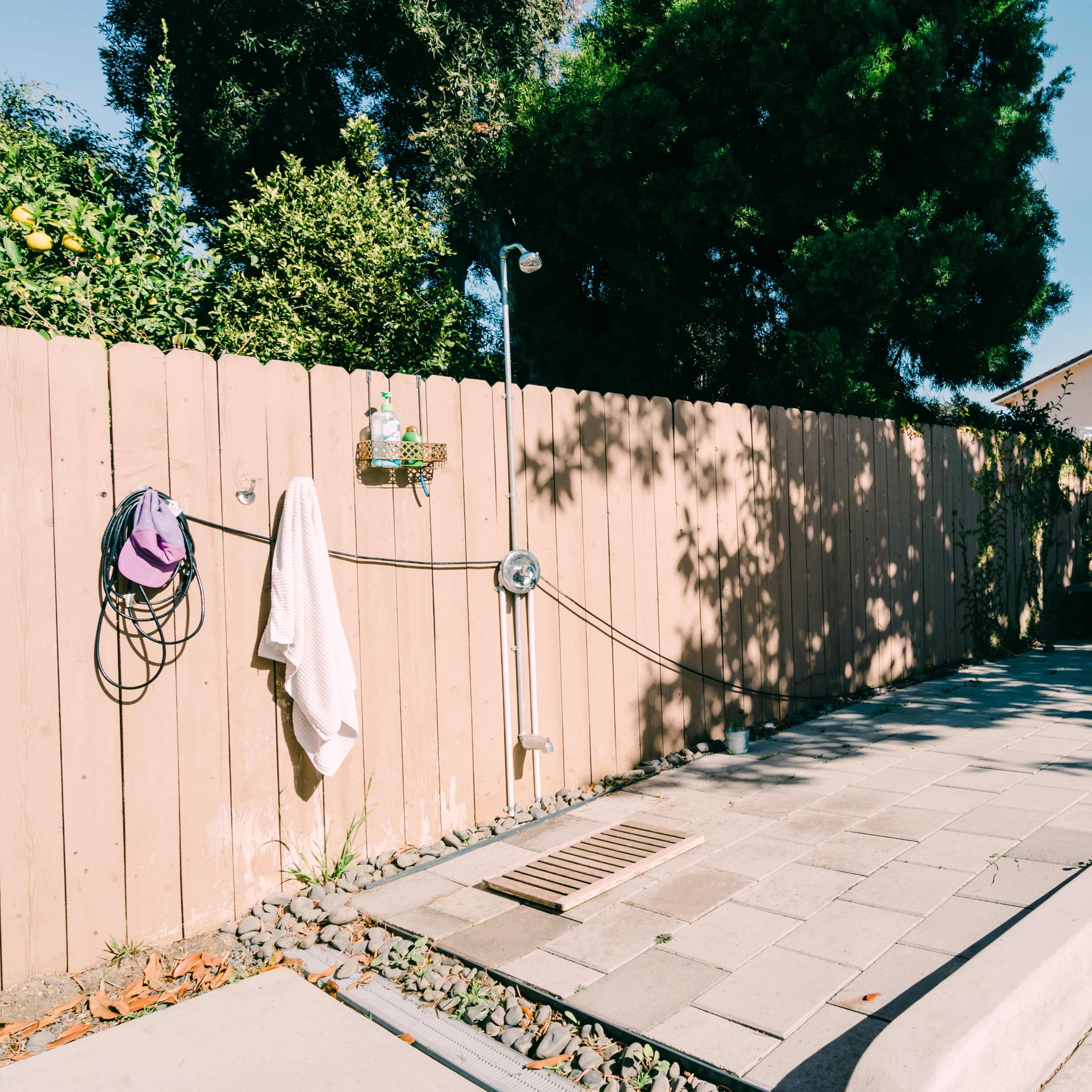 A sunny backyard features a paved area with a drainage grate, a hose connection, and a lounge chair next to a wooden fence.