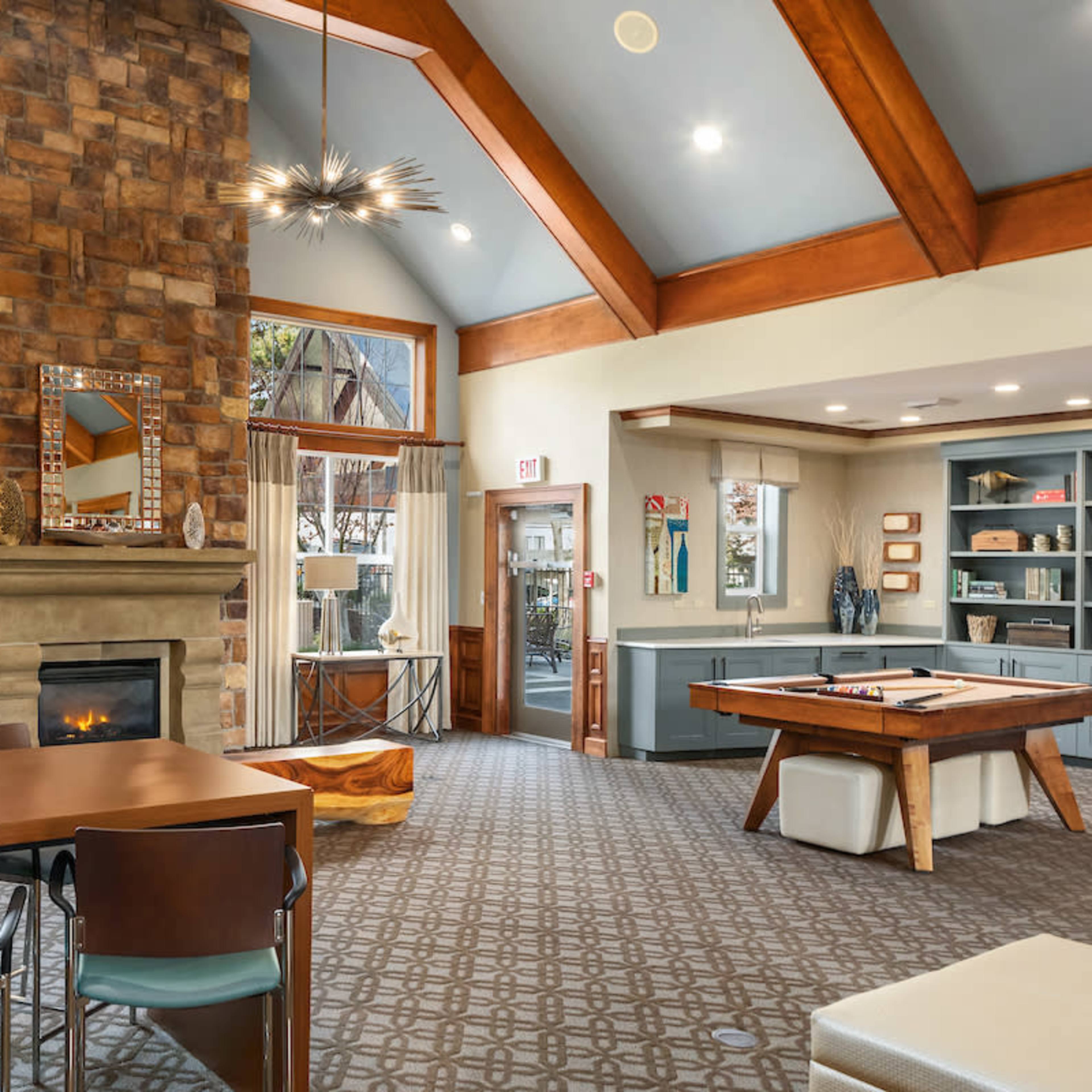 A modern interior space featuring a stone fireplace, wooden beams, a dining table, and a pool table, with large windows allowing natural light.