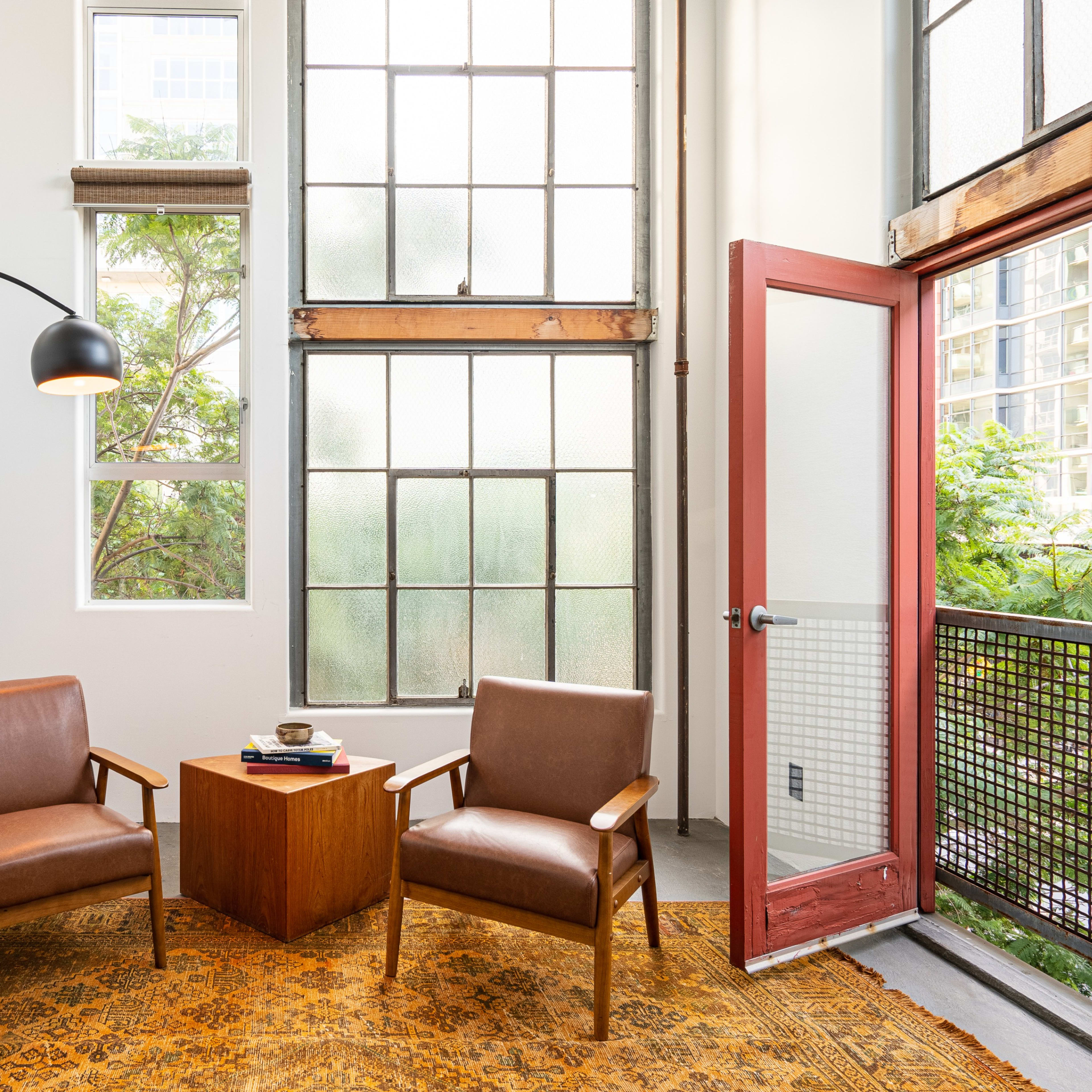 A seating area features two brown chairs beside a wooden side table, illuminated by a floor lamp, with large windows letting in natural light.