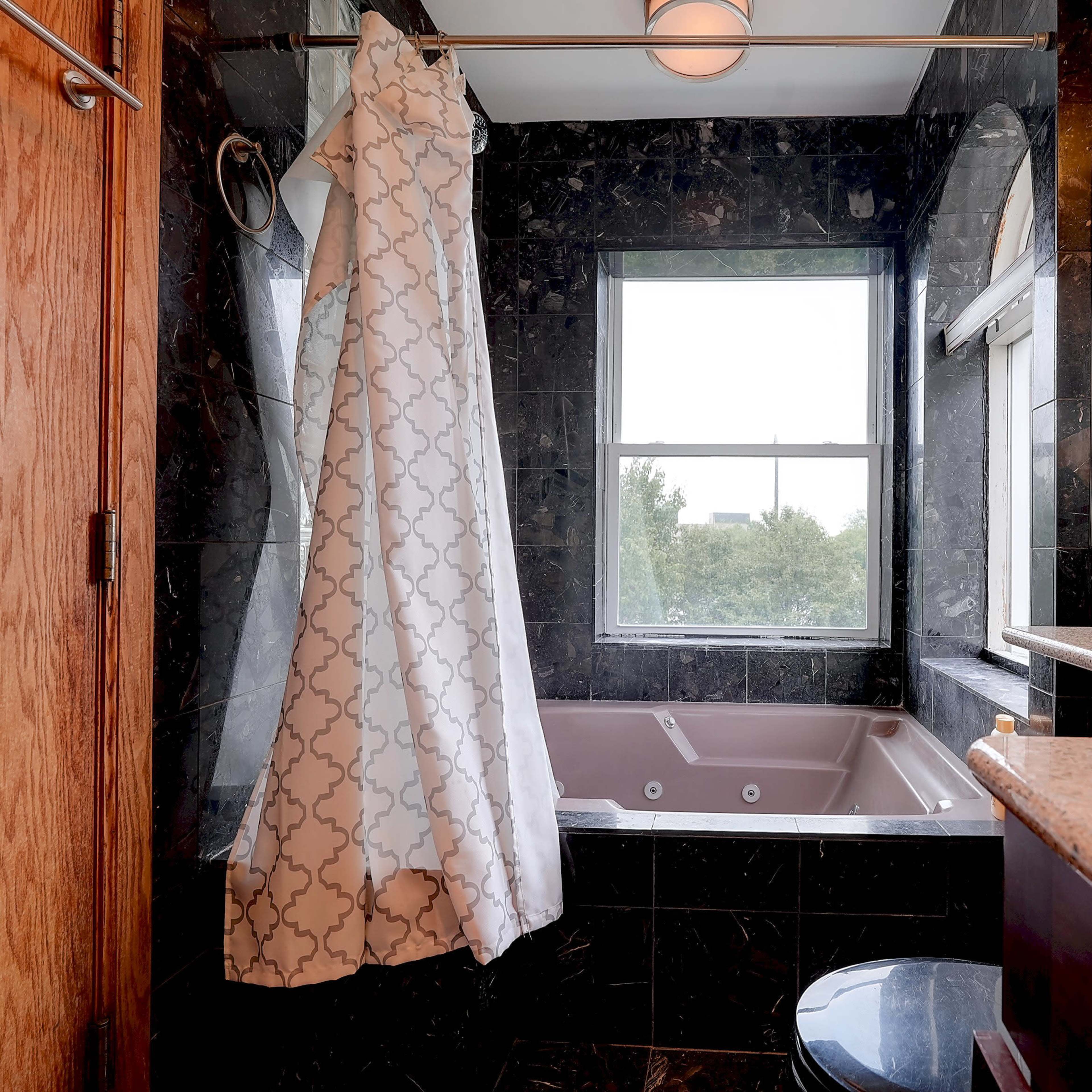 A bathroom featuring a bathtub with a shower curtain, a sink with a granite countertop, and large windows with a view of greenery outside.