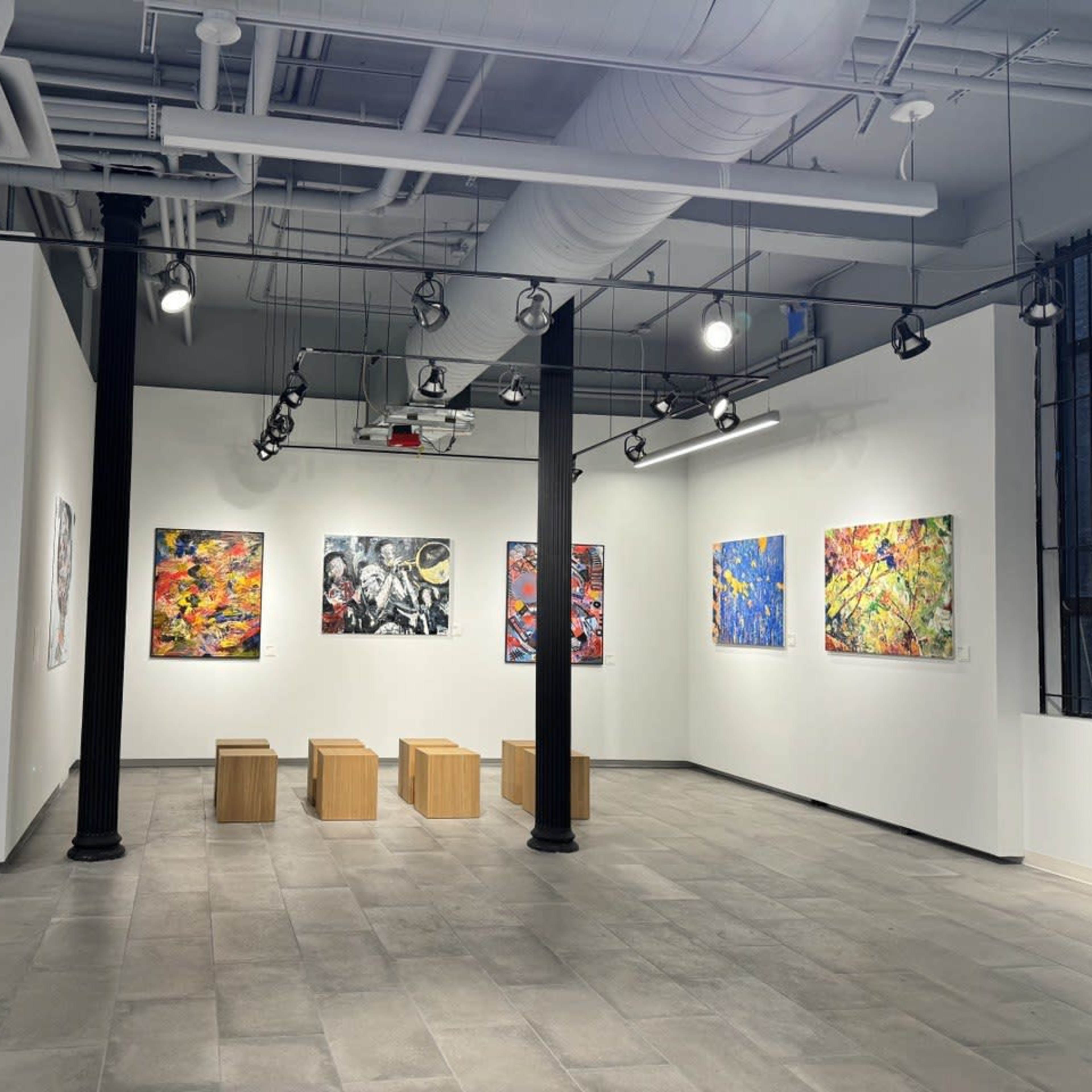 An art gallery featuring several colorful paintings on white walls, with wooden benches placed in the center.