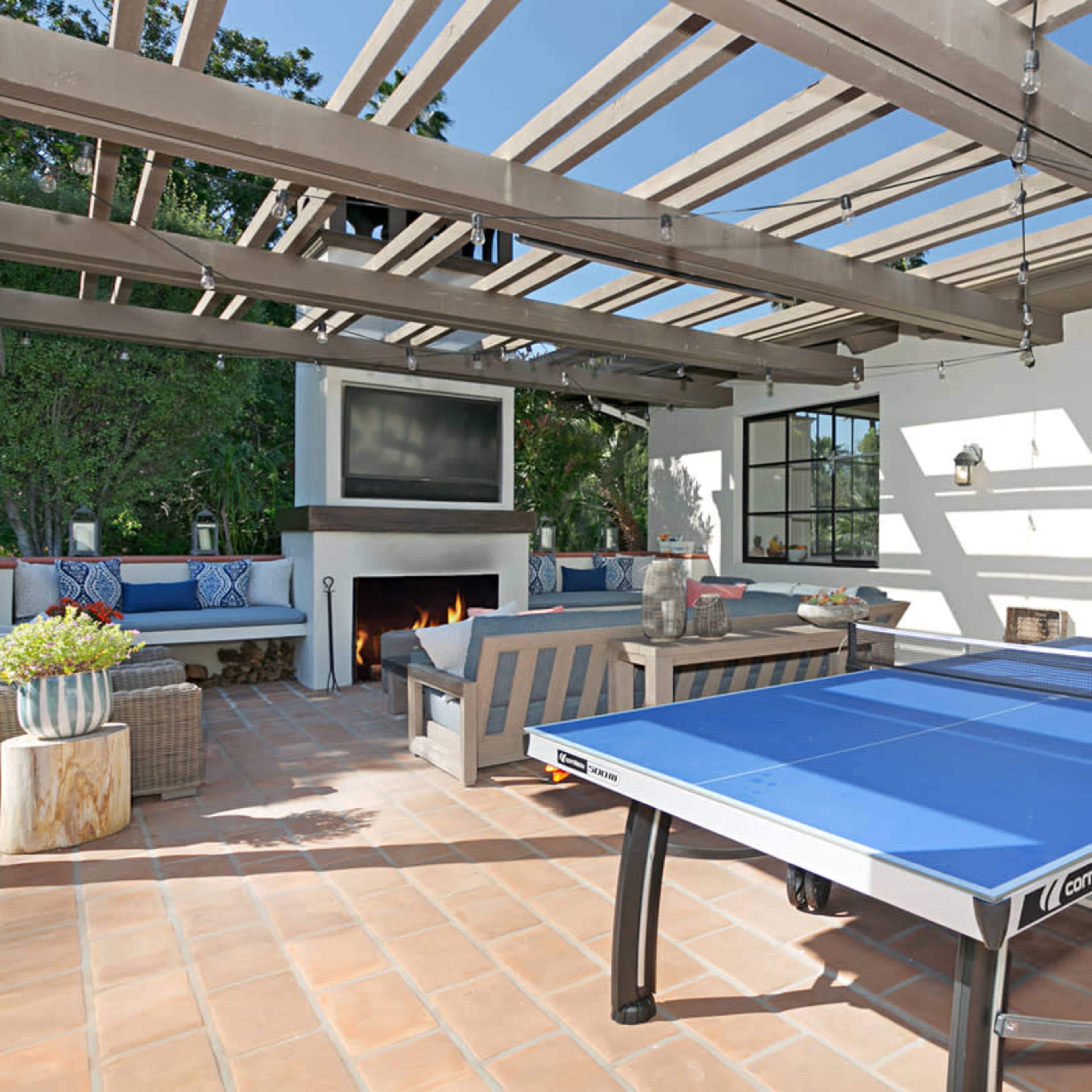 A patio features a blue ping pong table, comfortable seating, and a television mounted on the wall, all under a pergola.