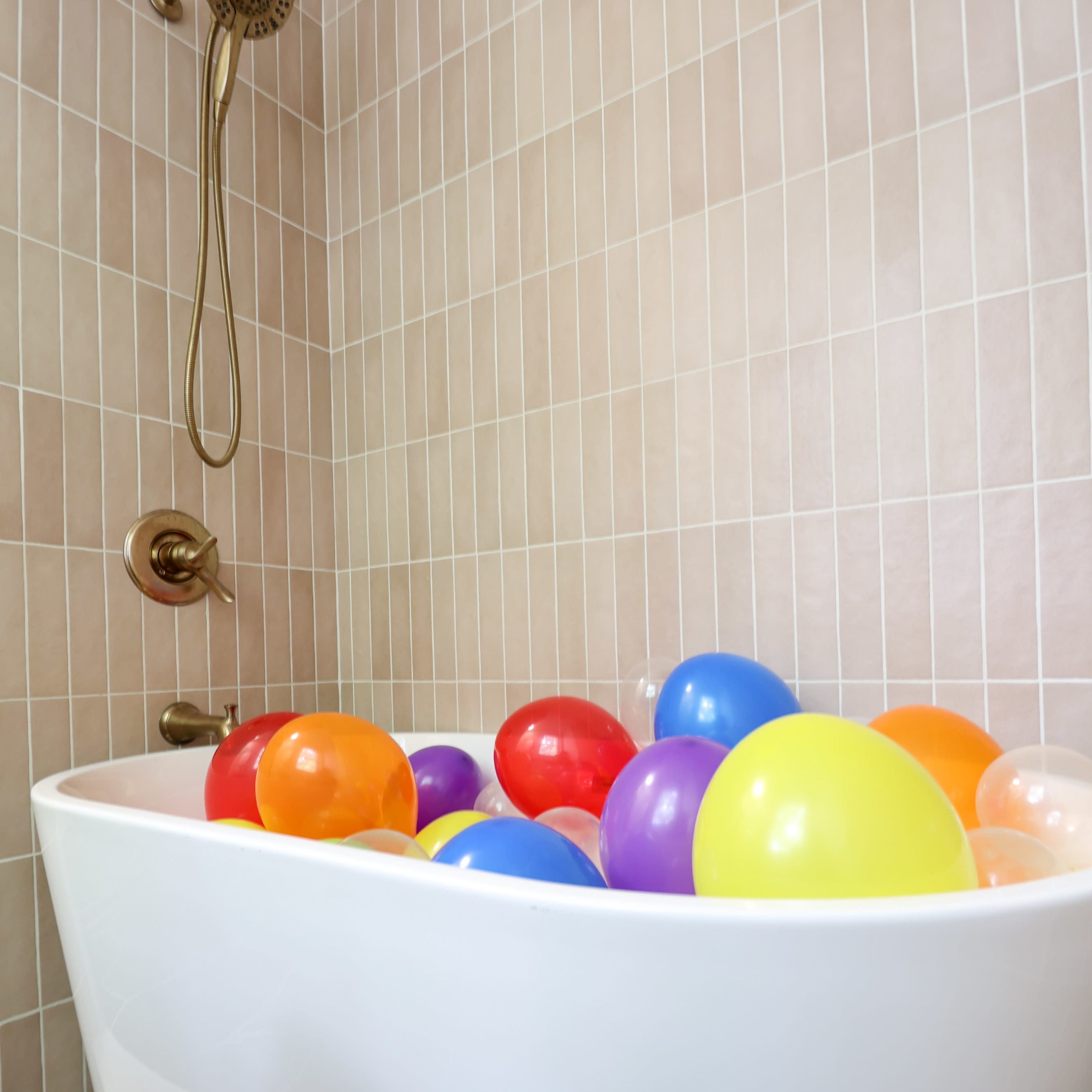 A white bathtub filled with colorful balloons sits in a tiled bathroom.