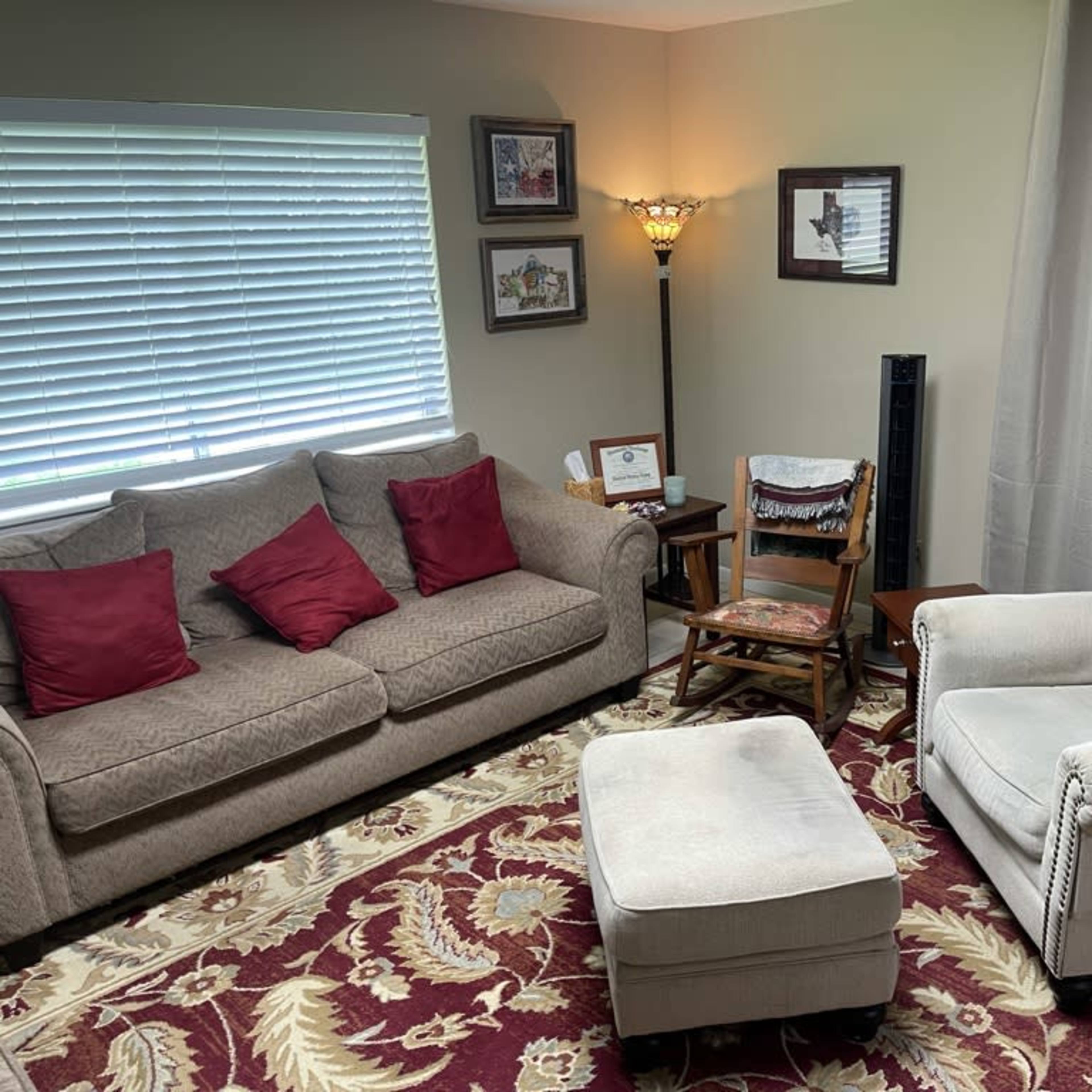 A cozy seating area with a gray sofa, an armchair, an ottoman, a wooden chair, and framed pictures on the wall, all arranged on a patterned rug.