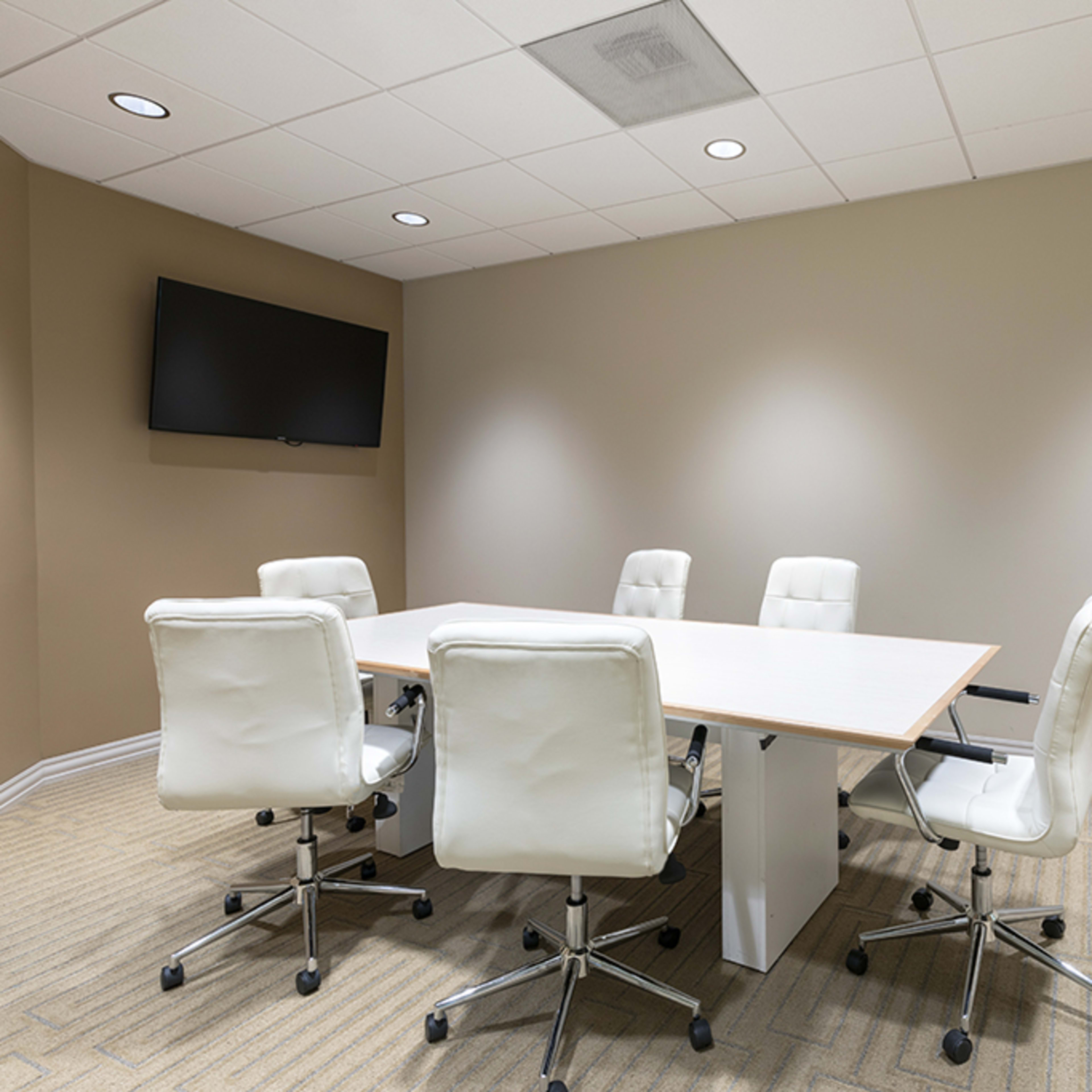A modern conference room features a large white table surrounded by five white chairs and a wall-mounted television.
