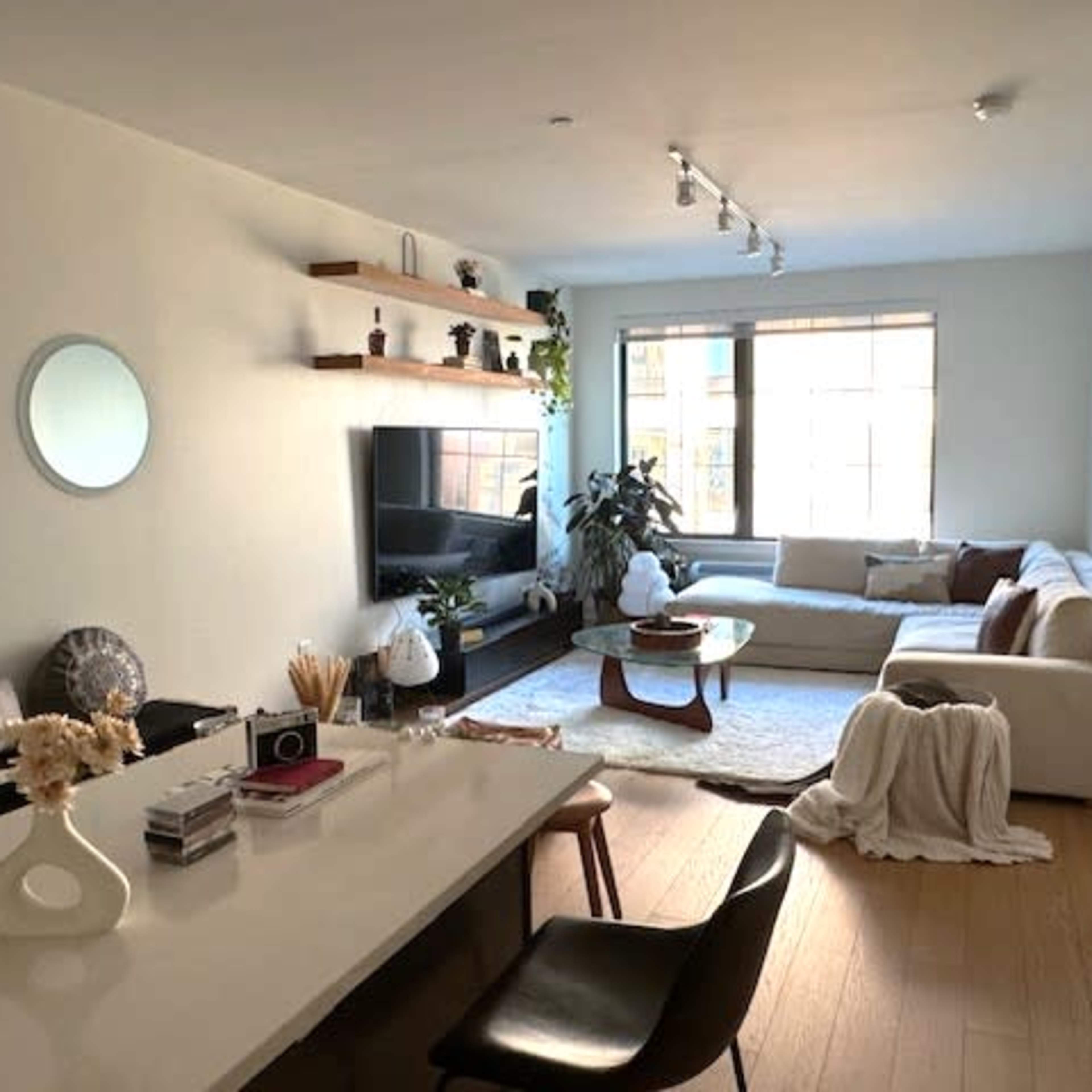 A modern living room with a sectional sofa, a coffee table, and a television mounted on the wall, alongside a kitchen area with sleek cabinetry and minimalist decor.